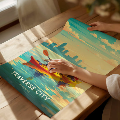 A person unrolls a vibrant Traverse City poster on a wooden table, depicting a kayaker on colorful waters under a sky with fluffy clouds.