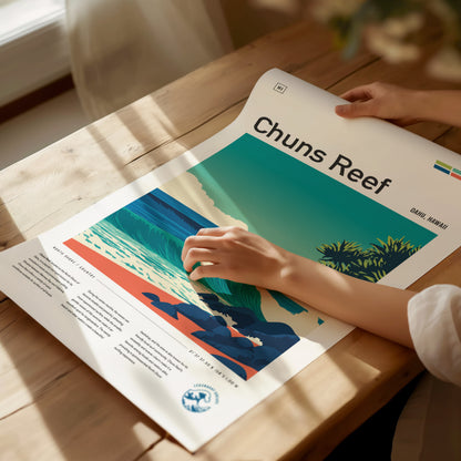 A person’s hand gently touches a vibrant poster featuring Chuns Reef, showcasing stylized waves and a coastal scene. The poster is laid on a wooden table, with natural light illuminating the artwork and surrounding greenery.