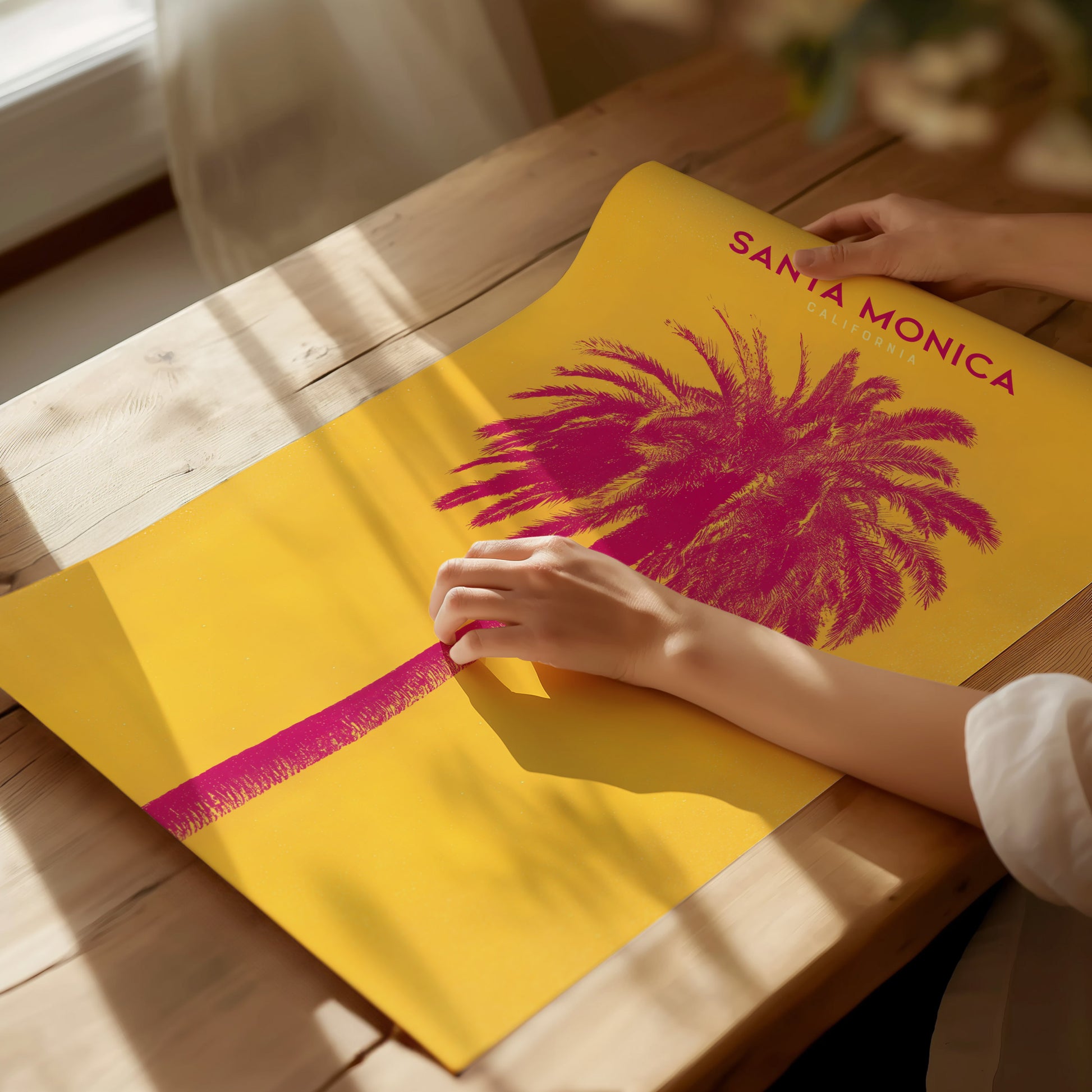 A person holds a vibrant poster featuring a pink palm tree against a yellow background with Santa Monica text. Sunlight casts soft shadows on a wooden table.