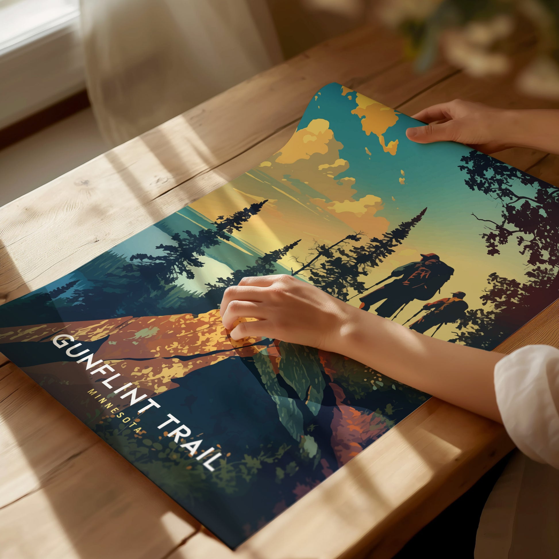 A person unrolls a colorful Gunflint Trail poster on a wooden table, featuring a scenic landscape with trees and hikers silhouetted against a vibrant sky.