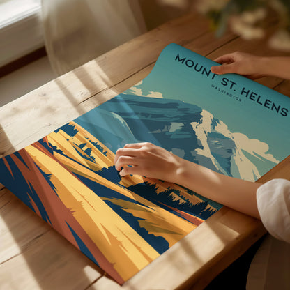 A person unrolls a colorful Mount St. Helens poster on a wooden table, depicting vibrant landscapes with blue mountains.