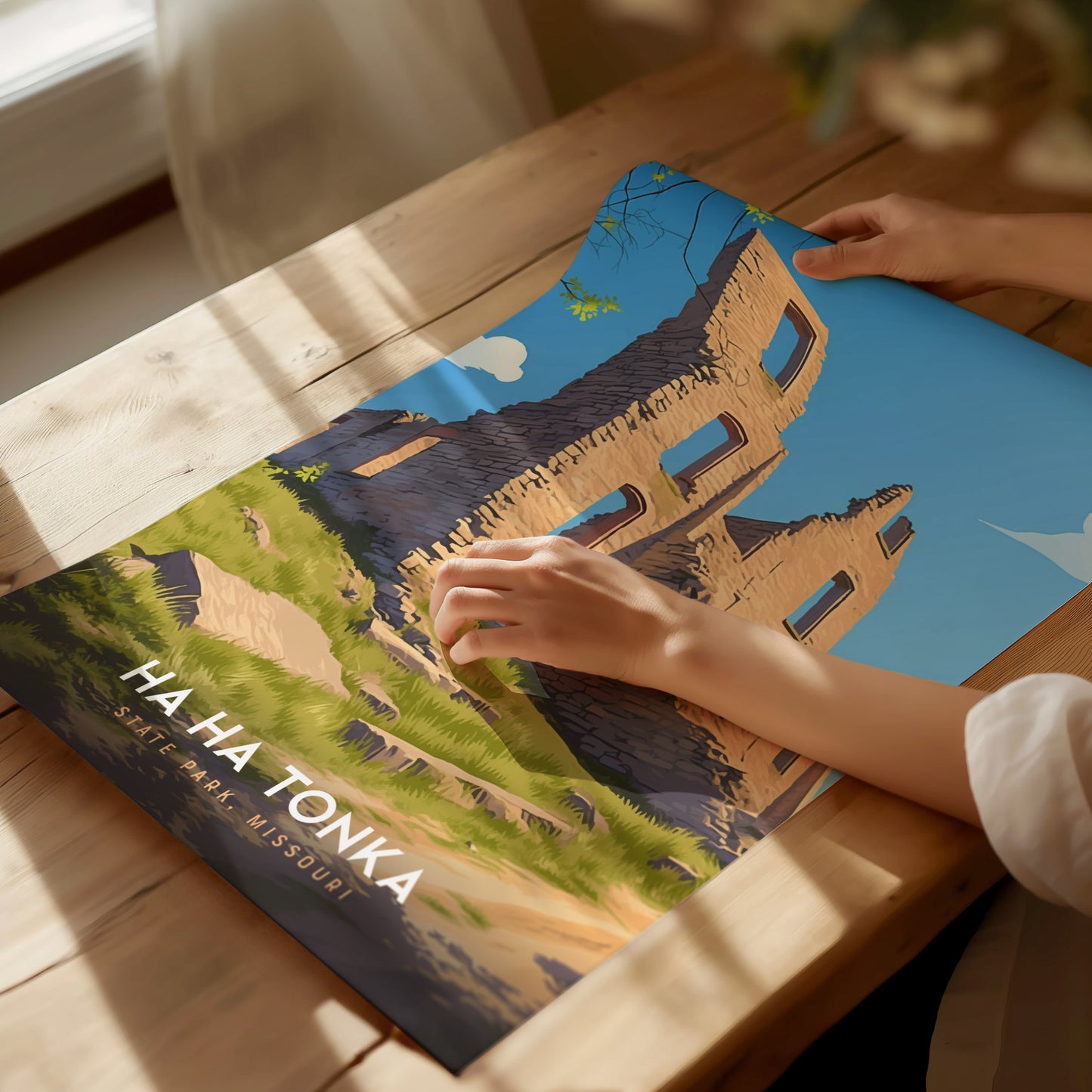 A person examines a vibrant poster depicting Ha Ha Tonka State Park, Missouri, showing a stone structure against a clear blue sky. The poster is laid on a wooden table.
