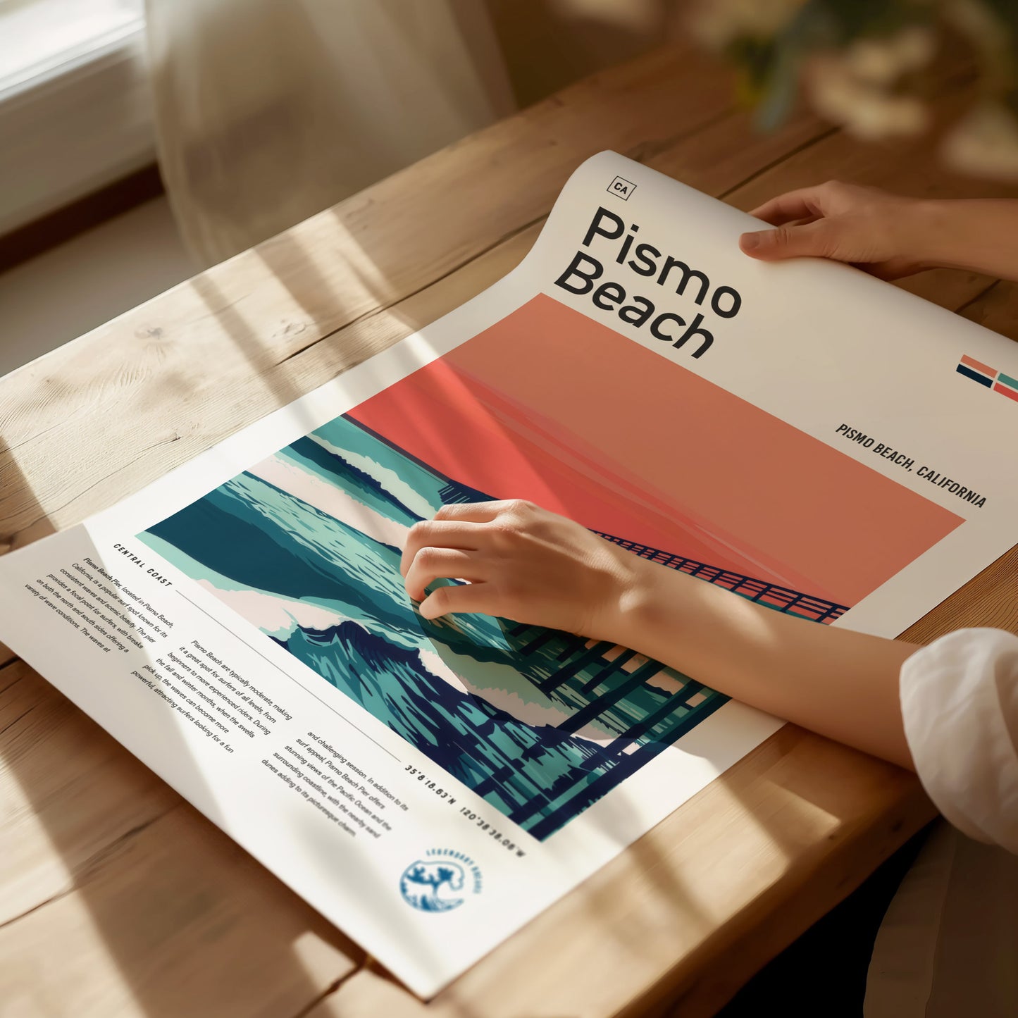 A person holds a Pismo Beach poster featuring a stylized depiction of the pier and ocean waves against a reddish-orange sky, laid on a wooden table.