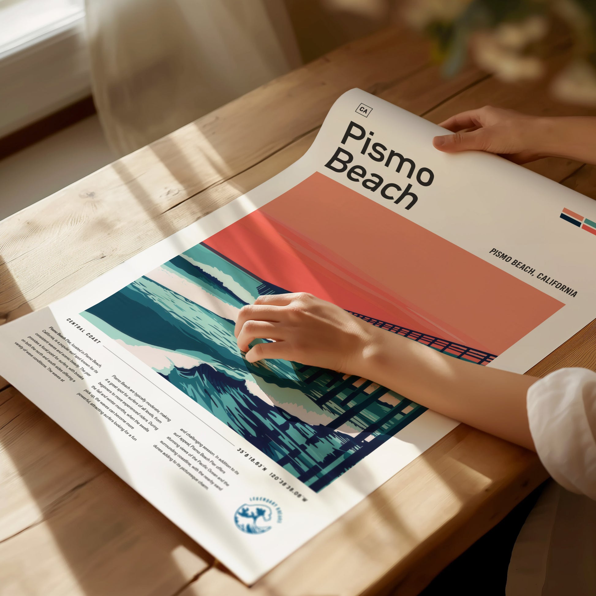 A person holds a Pismo Beach poster featuring a stylized depiction of the pier and ocean waves against a reddish-orange sky, laid on a wooden table.