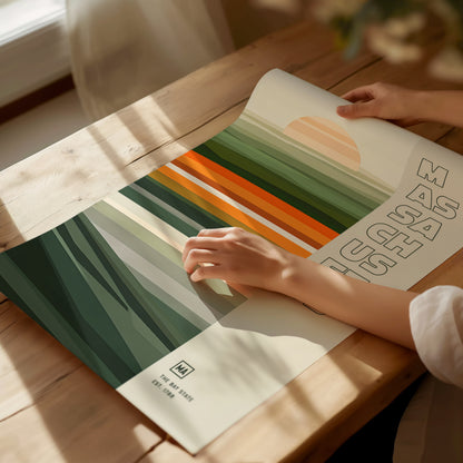 A person adjusts a colorful, striped poster on a wooden table with sunlight streaming in. The poster features green and orange tones with abstract lettering on the right.