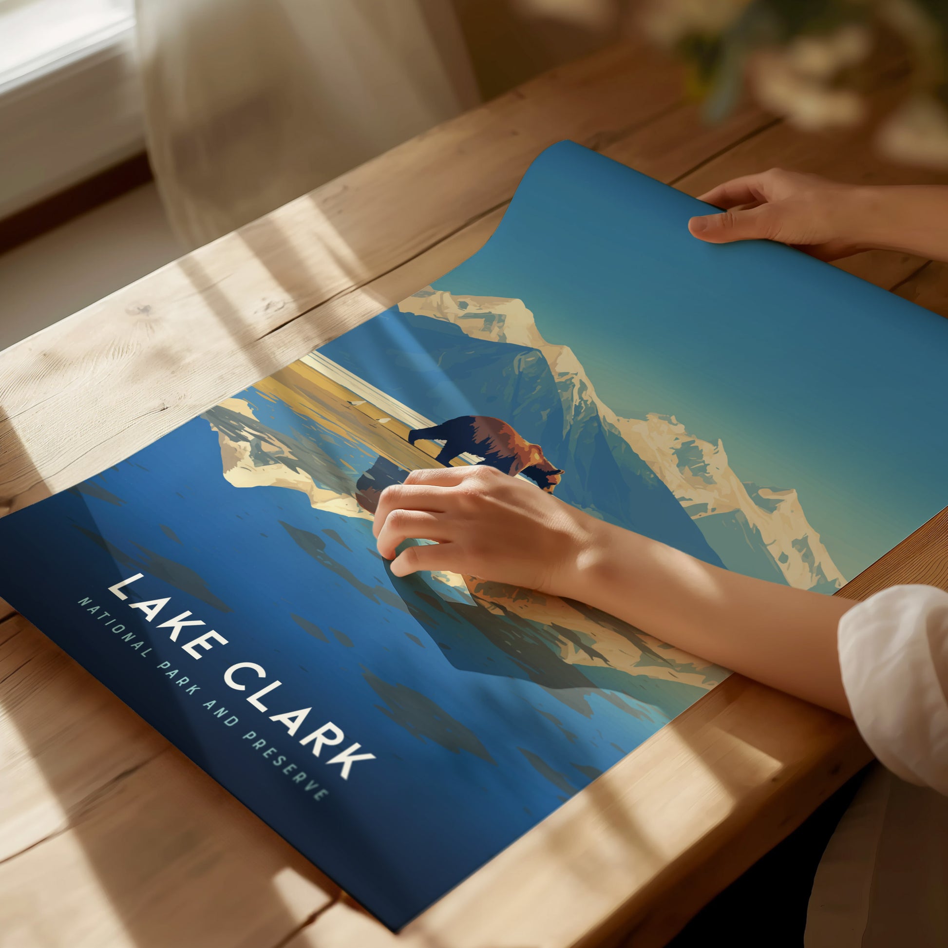 A person unrolls a vibrant Lake Clark National Park poster featuring mountains and a bear on a wooden table. Sunlight casts soft shadows across the scene.