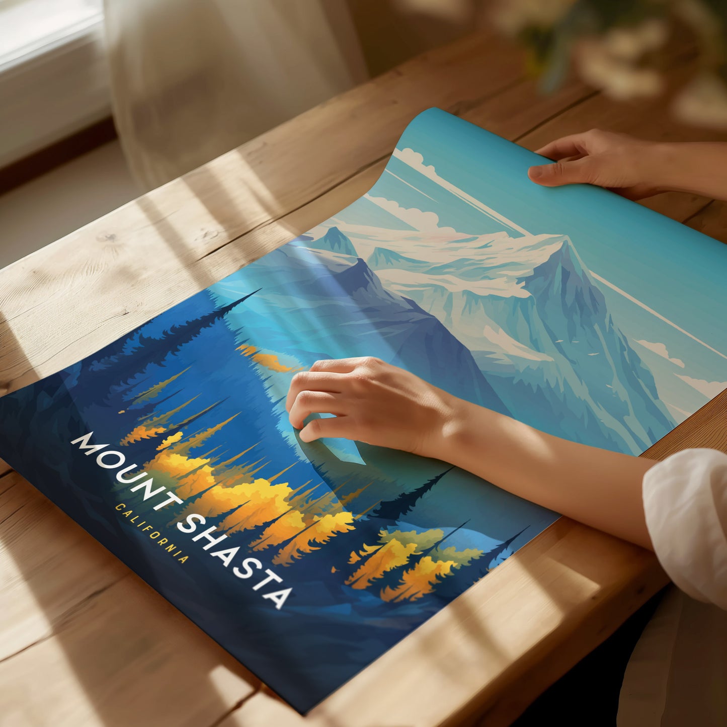 A person unrolls a vibrant Mount Shasta poster on a wooden table, featuring snowy peaks and colorful forested foothills under a clear blue sky. Sunlight streams through a nearby window.