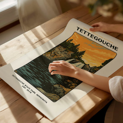 A person arranges a Tettegouche State Park poster on a wooden table, showing a scenic illustration of cliffs, trees, and a waterfall under an orange sky.