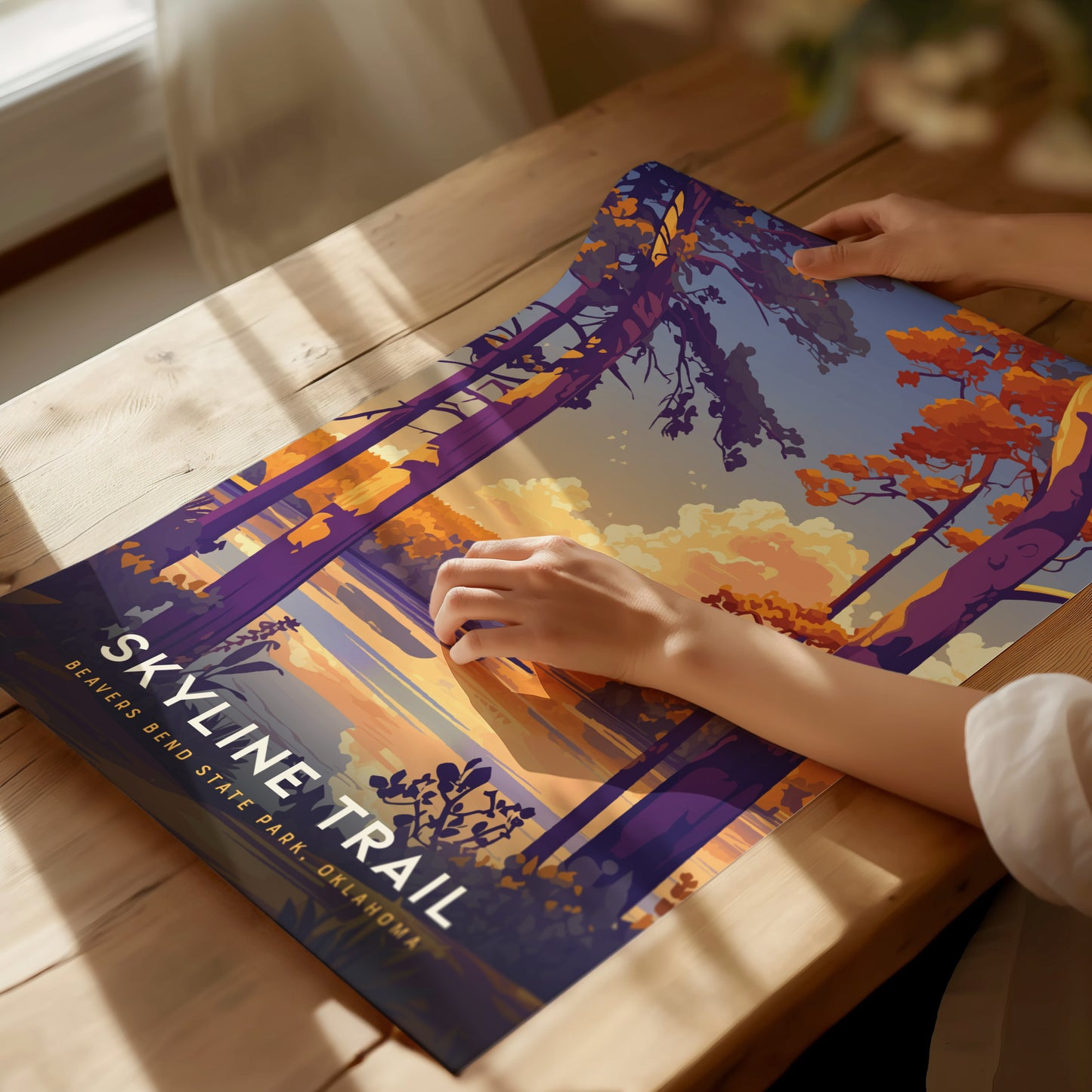 A person places a colorful poster of the Skyline Trail at Beavers Bend State Park on a wooden table. The artwork features vibrant trees and a scenic landscape.