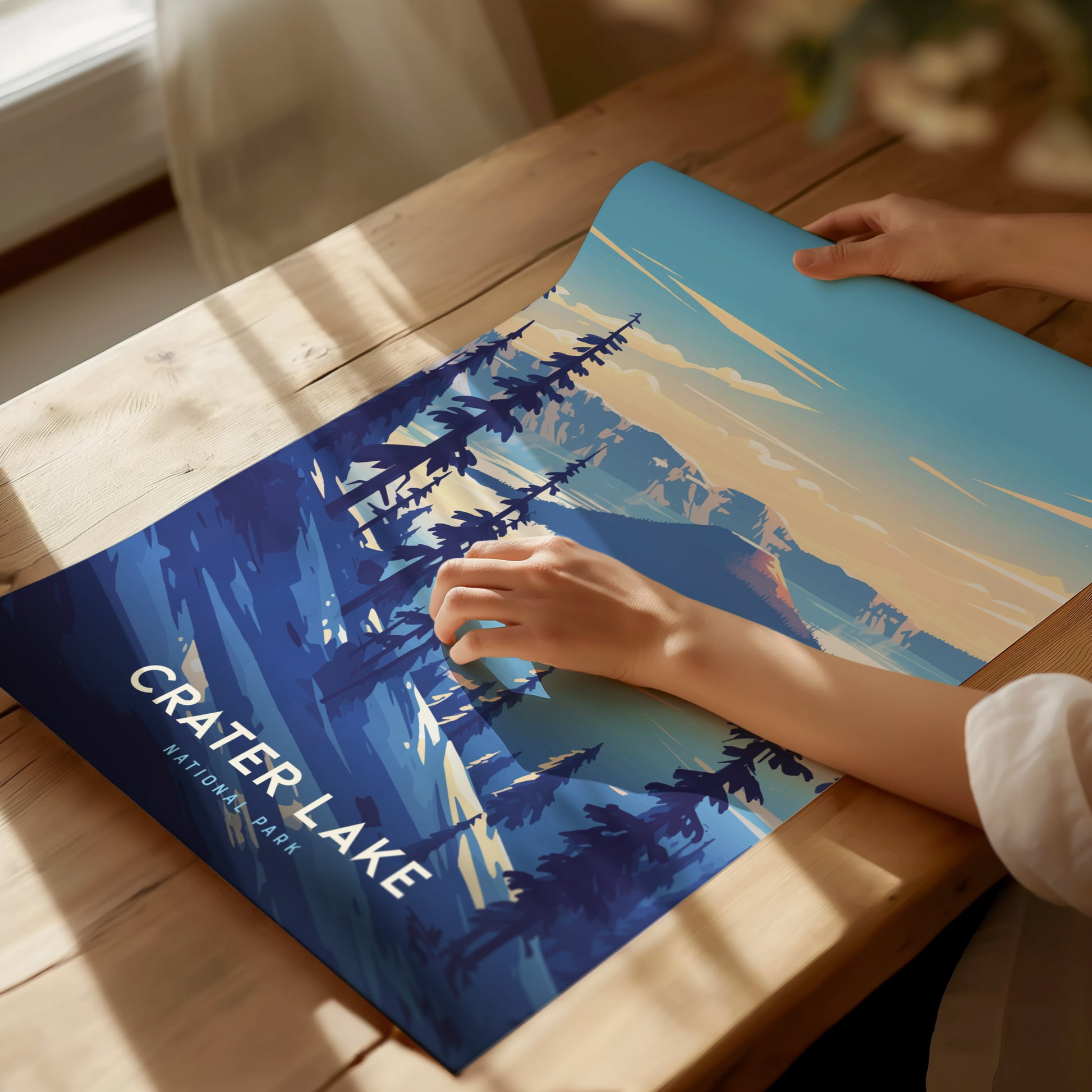 A person unrolls a vibrant Crater Lake National Park poster on a wooden table, featuring stylized blue and orange landscapes with silhouetted trees. Sunlight casts shadows across the scene.