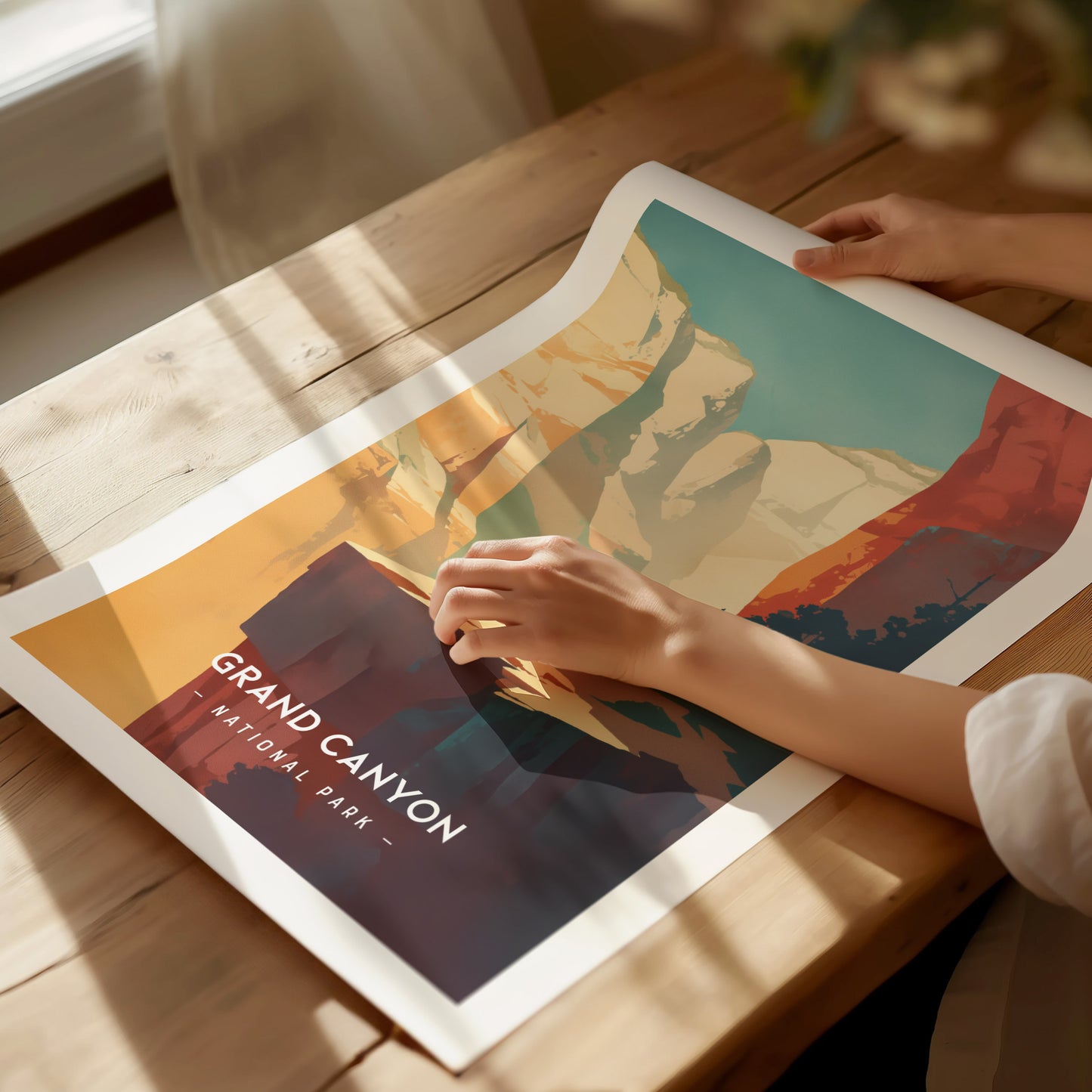A person arranges a colorful Grand Canyon National Park poster on a wooden table. Sunlight casts soft shadows across the minimalistic illustration with vibrant hues and text.