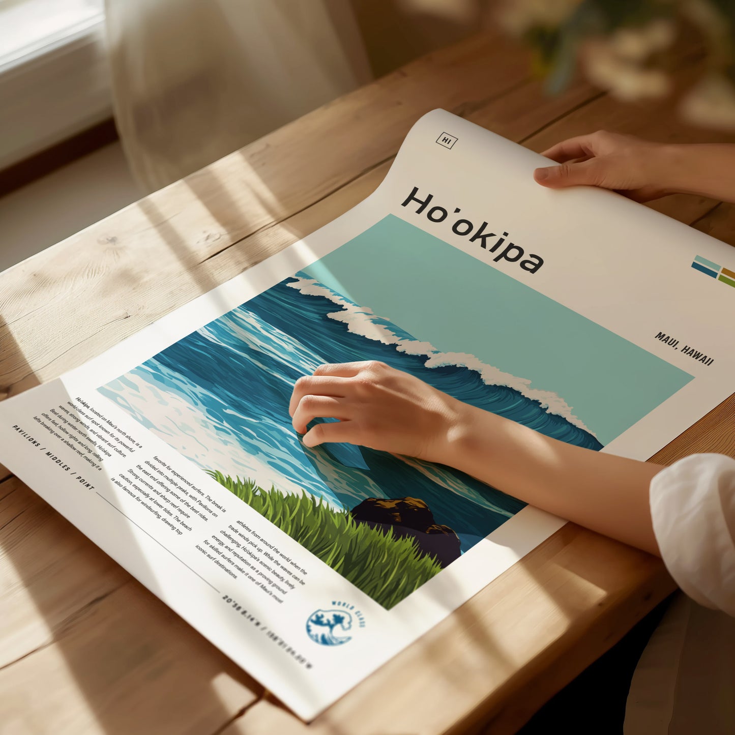 A person gently holds a Hookipa Beach poster featuring ocean waves and grassy cliffs, displayed on a wooden table with soft natural lighting. The poster highlights Maui, Hawaii.