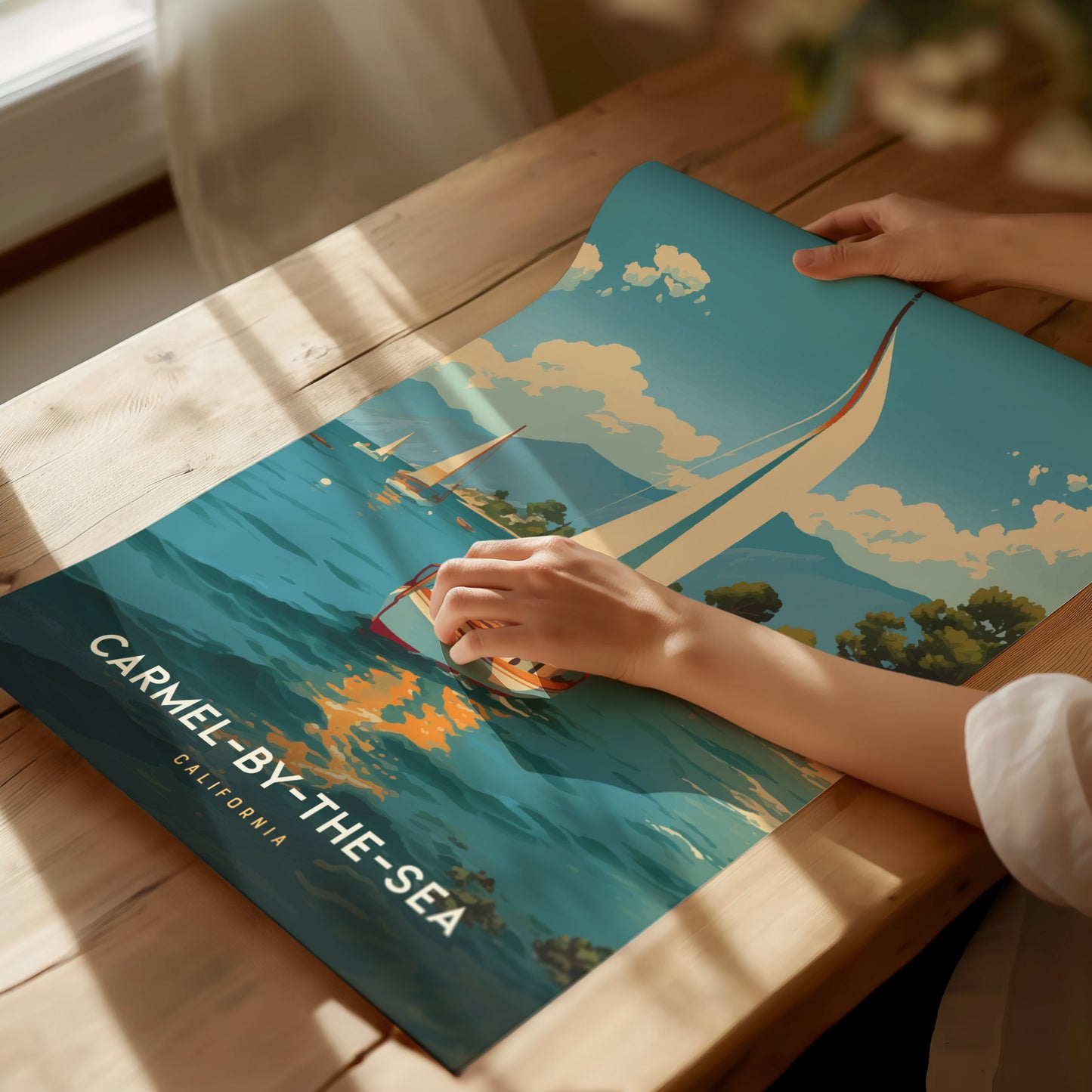 A person holds a colorful poster depicting a sailboat on the ocean with the text Carmel-by-the-Sea, California on a wooden table. Sunlight streams through a nearby window.