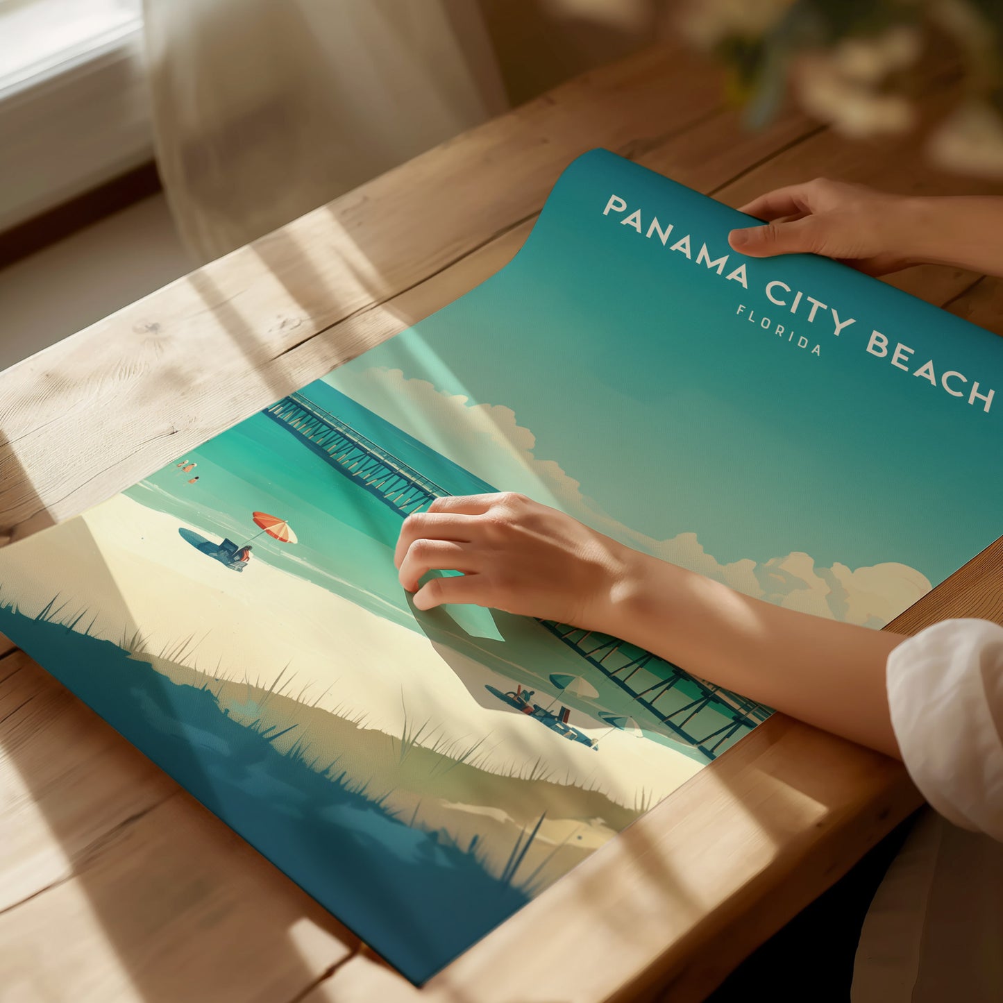 A person unrolls a vibrant Panama City Beach poster on a wooden table, featuring turquoise waters, a sandy beach, and a pier under a sunny sky.