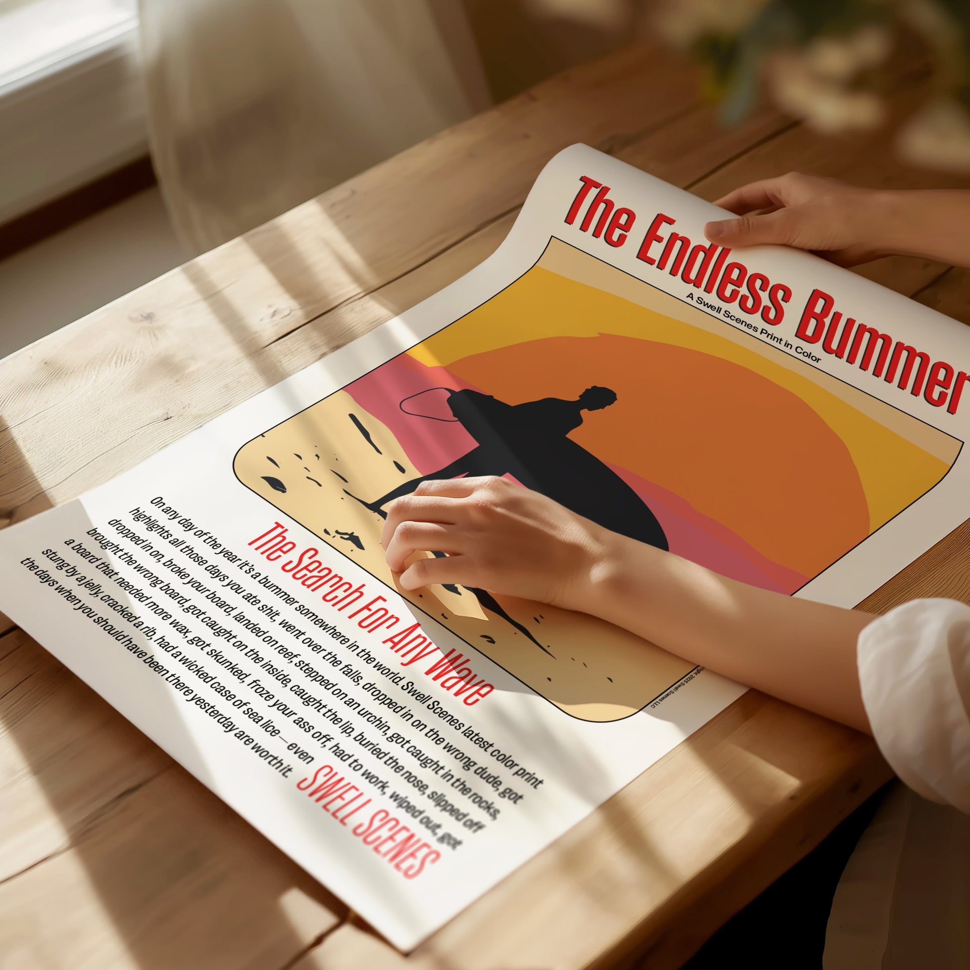 A person unrolls a poster titled The Endless Bummer on a wooden table, featuring a silhouetted surfer against an orange sunset. Sunlight streams through a window onto the scene.