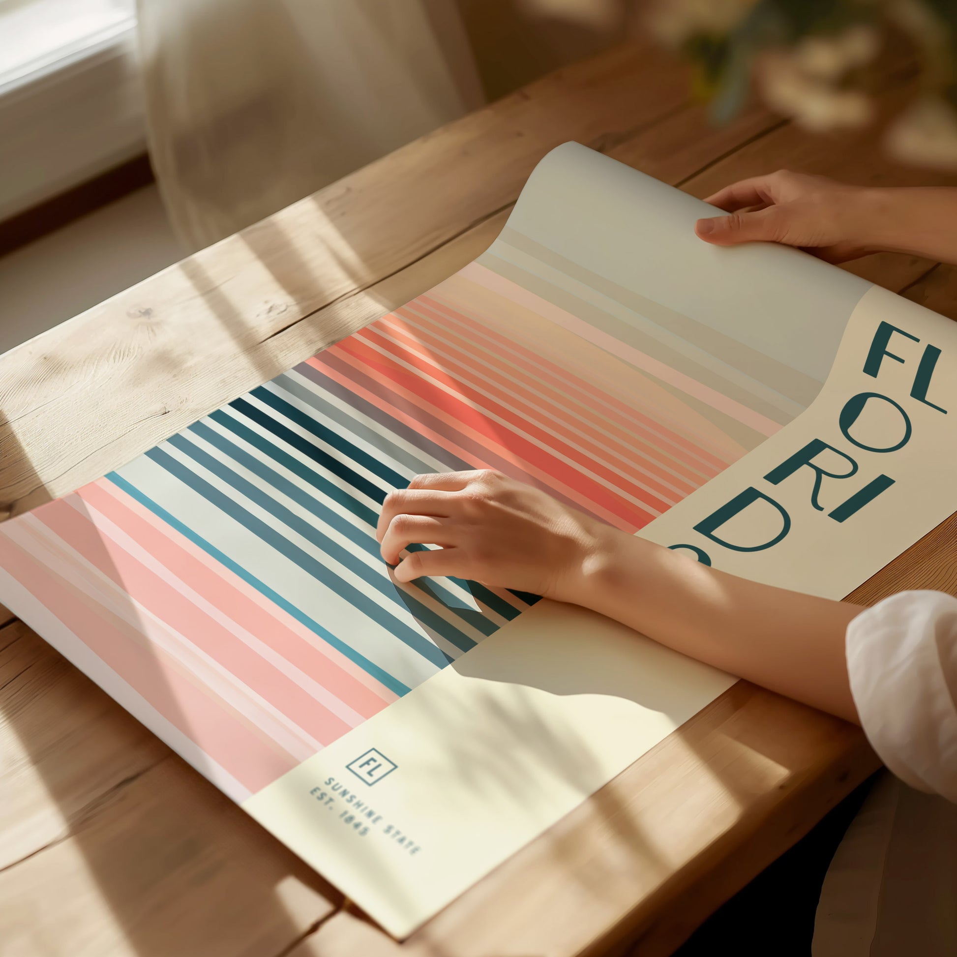A person arranges a colorful poster on a wooden table, featuring vertical stripes in pastel shades and the word FLORIDA in bold letters. Sunlight casts gentle shadows across the scene.