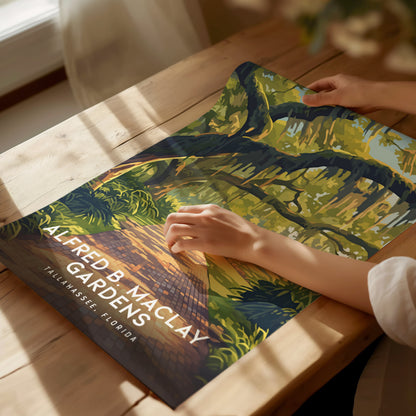 A person gently holds an illustrated poster of Alfred B. Maclay Gardens, showcasing vibrant trees and a brick path. Sunlight streams through a nearby window onto the wooden table.