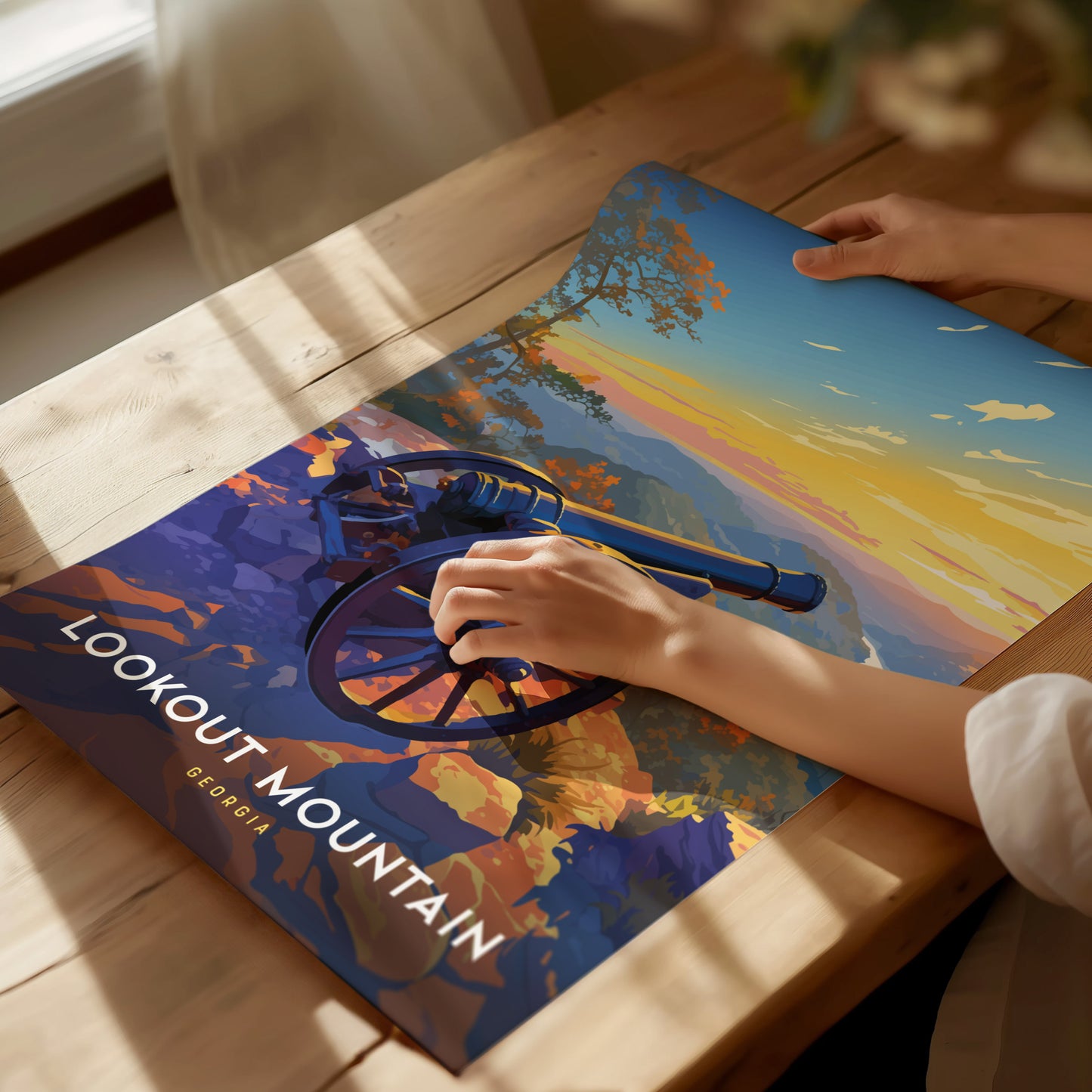 A person unrolls a colorful poster of Lookout Mountain, Georgia, featuring a cannon and a vibrant sunset landscape, on a wooden table. Sunlight streams through a nearby window.