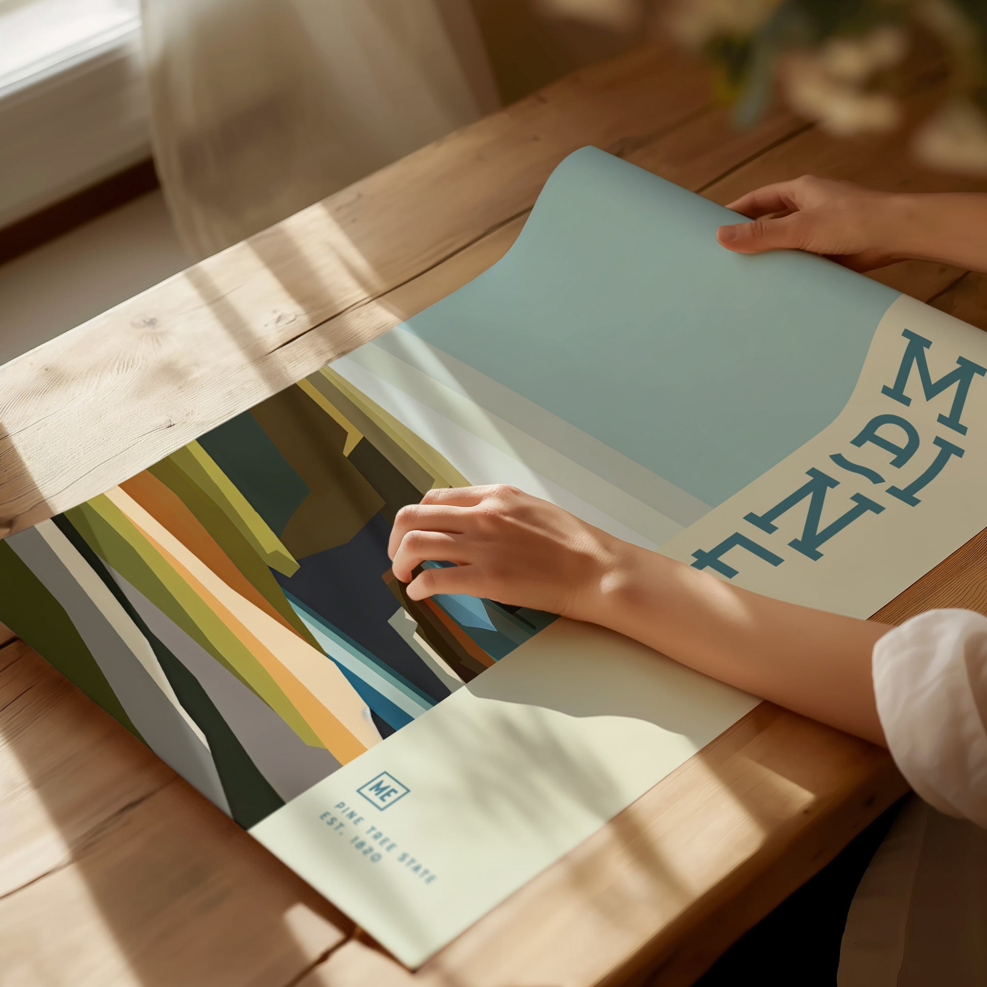 A person holds a colorful minimalist poster with abstract geometric shapes on a wooden table, sunlight streaming in from a window. The word MAINE is visible in blue letters.