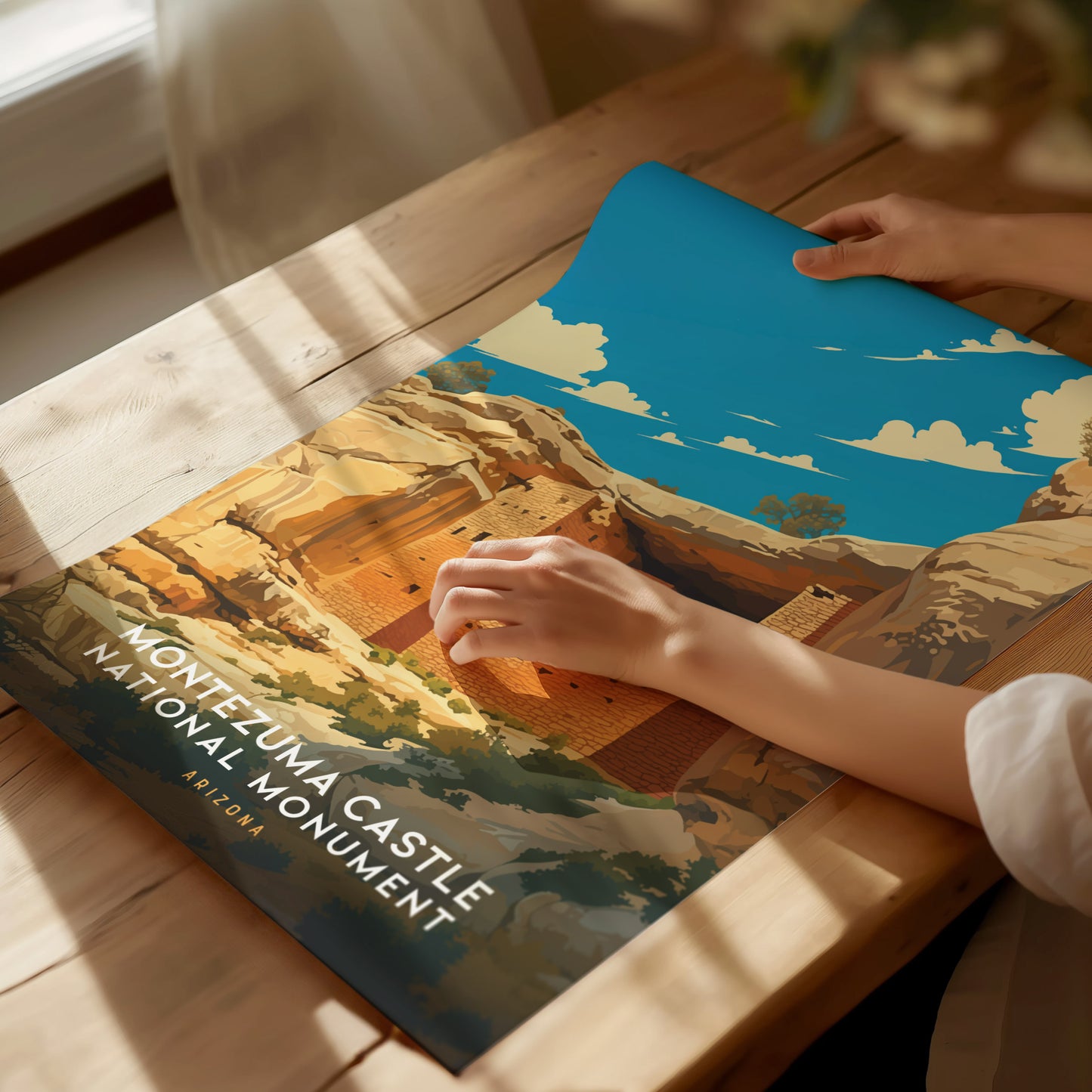 A person rolls out a colorful poster of Montezuma Castle National Monument on a wooden table. Sunlight streams through a window, highlighting the vibrant blue sky and desert cliffs.