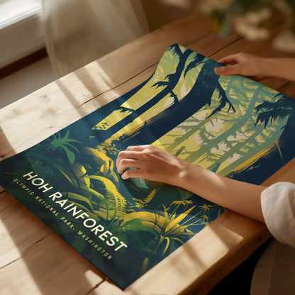 A person holds a colorful poster depicting the Hoh Rainforest, featuring vibrant green and yellow hues with trees and foliage. Sunlight casts shadows on the wooden table.