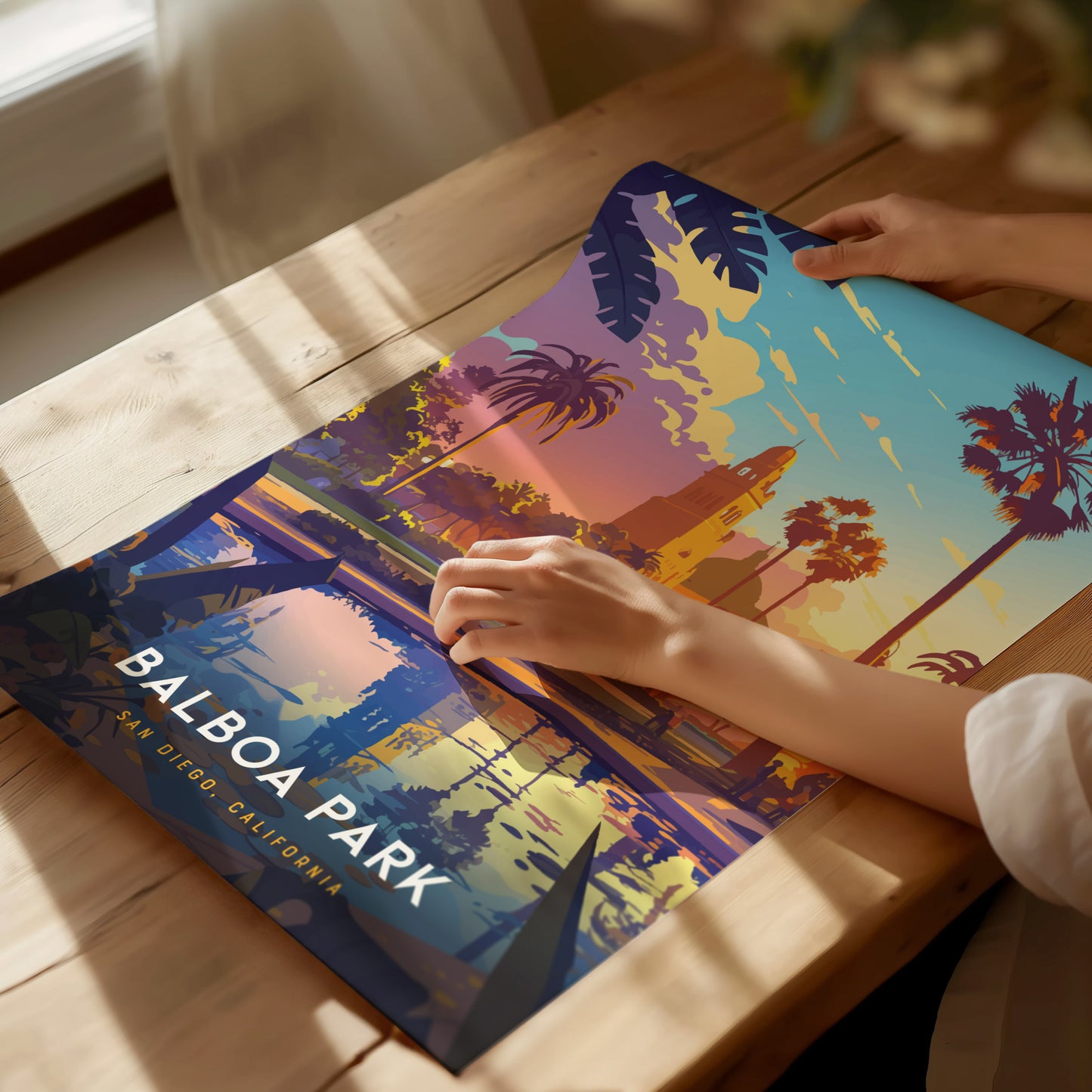 A person examines a colorful Balboa Park poster on a wooden table, showcasing vibrant palm trees and architecture under a sunset sky. Sunlight streams through a nearby window.