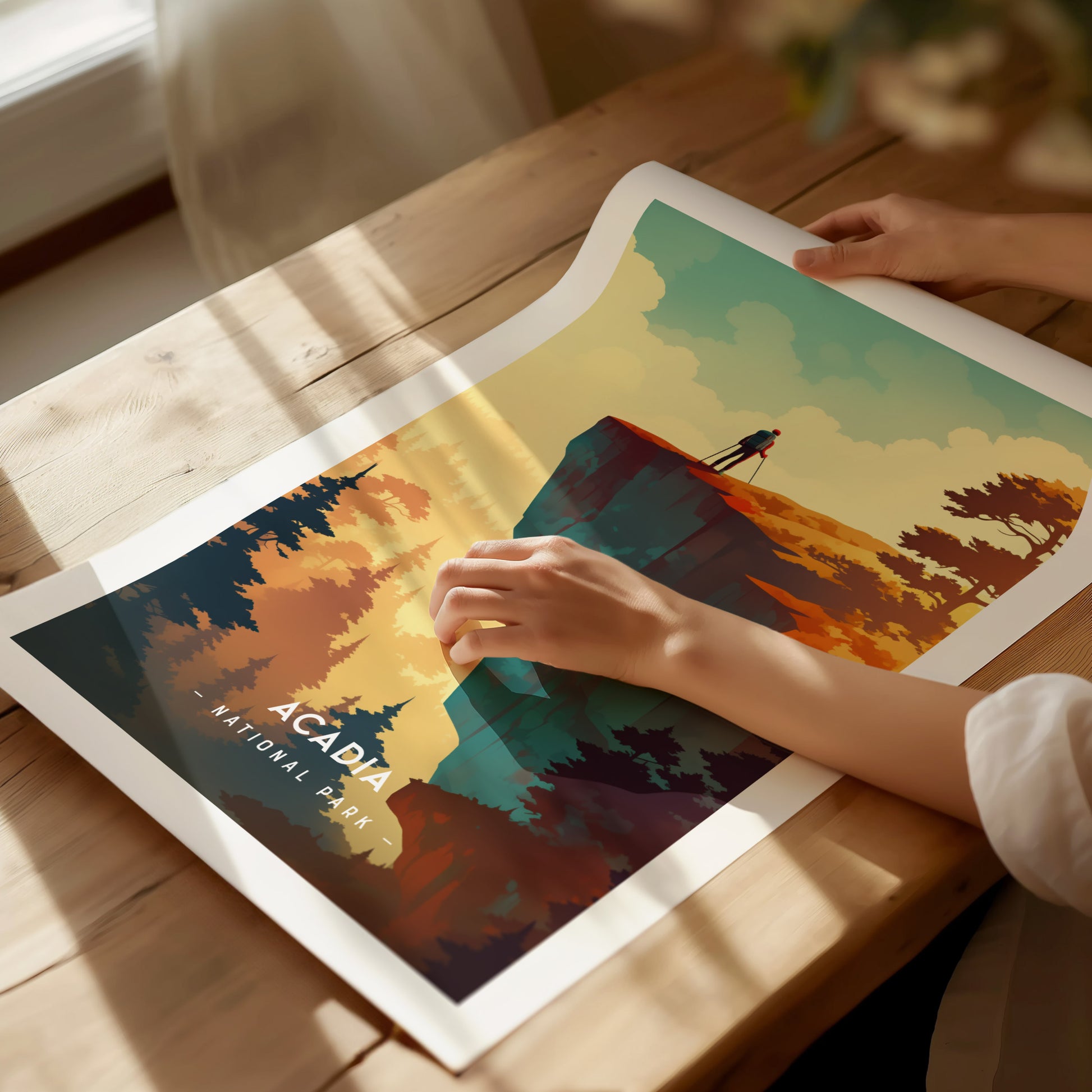 A person examines a colorful Acadia National Park poster featuring a lone figure on a cliff, surrounded by trees and a vibrant sky, on a sunlit wooden table.