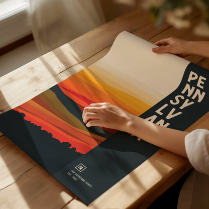 A person unrolls a colorful, abstract Pennsylvania poster on a sunlit wooden table, featuring layered shades of orange and yellow with bold text spelling PENNSYLVANIA.
