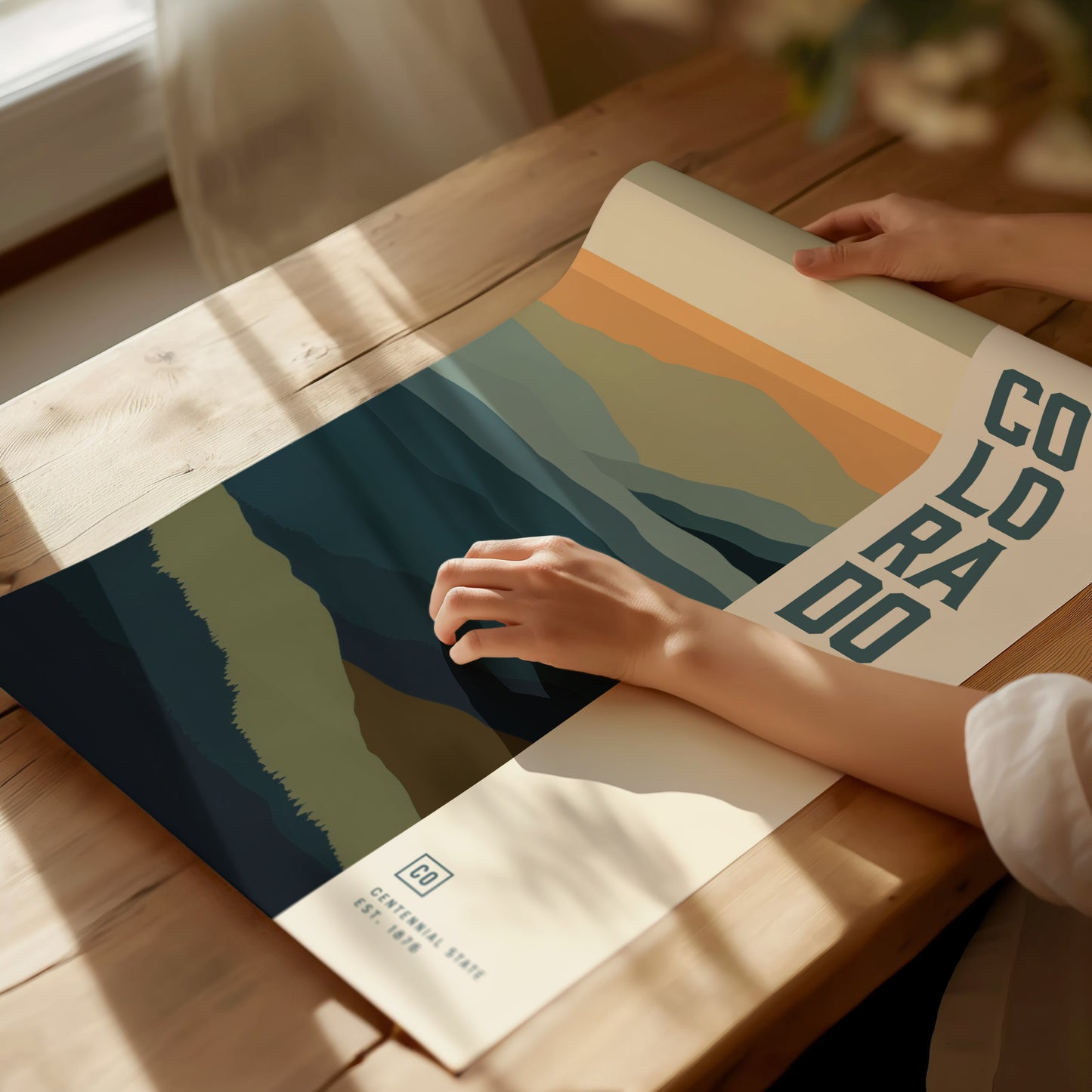 A person unrolls a minimalist Colorado poster featuring abstract mountain designs in soft hues on a wooden table. Sunlight casts gentle shadows across the scene, enhancing the serene atmosphere.