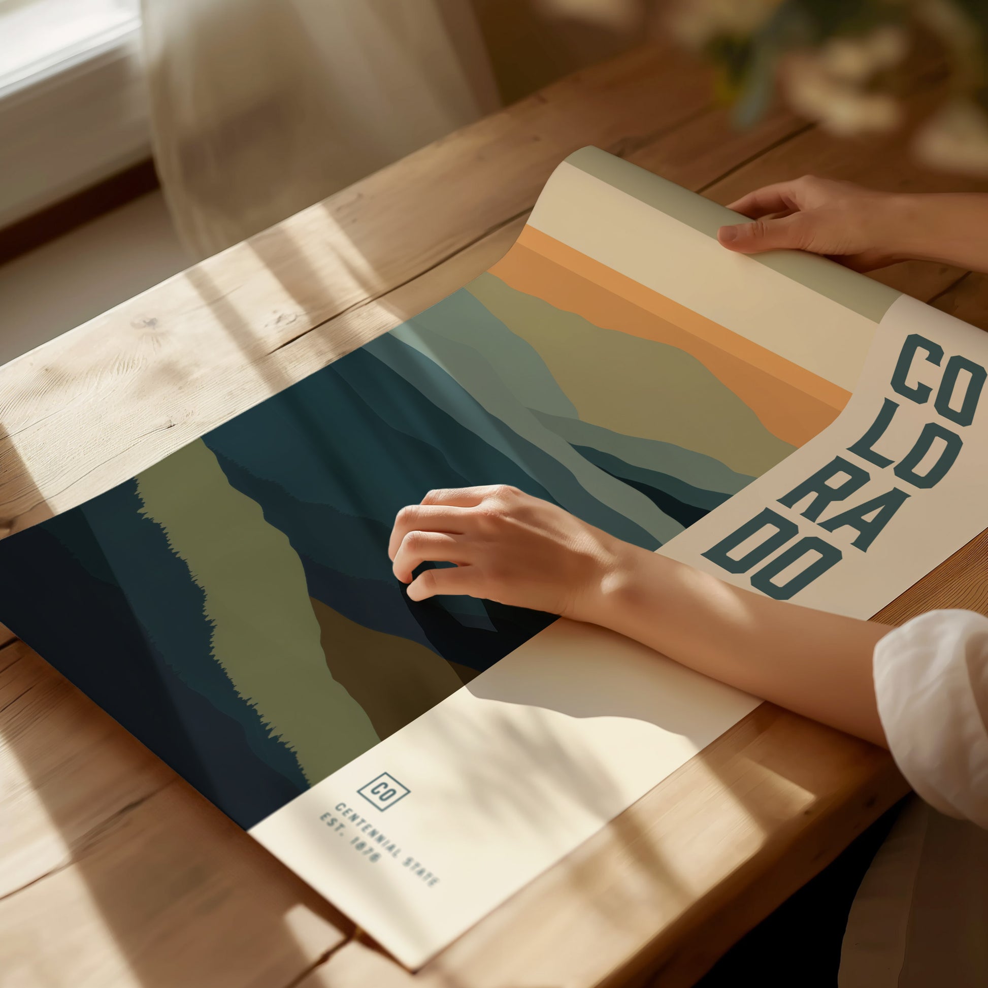 A person unrolls a minimalist Colorado poster featuring abstract mountain designs in soft hues on a wooden table. Sunlight casts gentle shadows across the scene, enhancing the serene atmosphere.