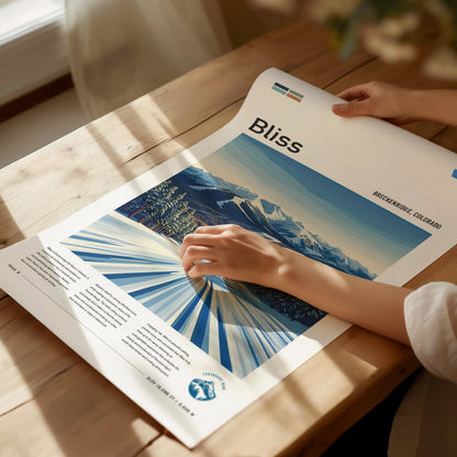 A person examines a Bliss ski run poster featuring stylized mountains and trees in Breckenridge, Colorado, on a wooden table in soft.
