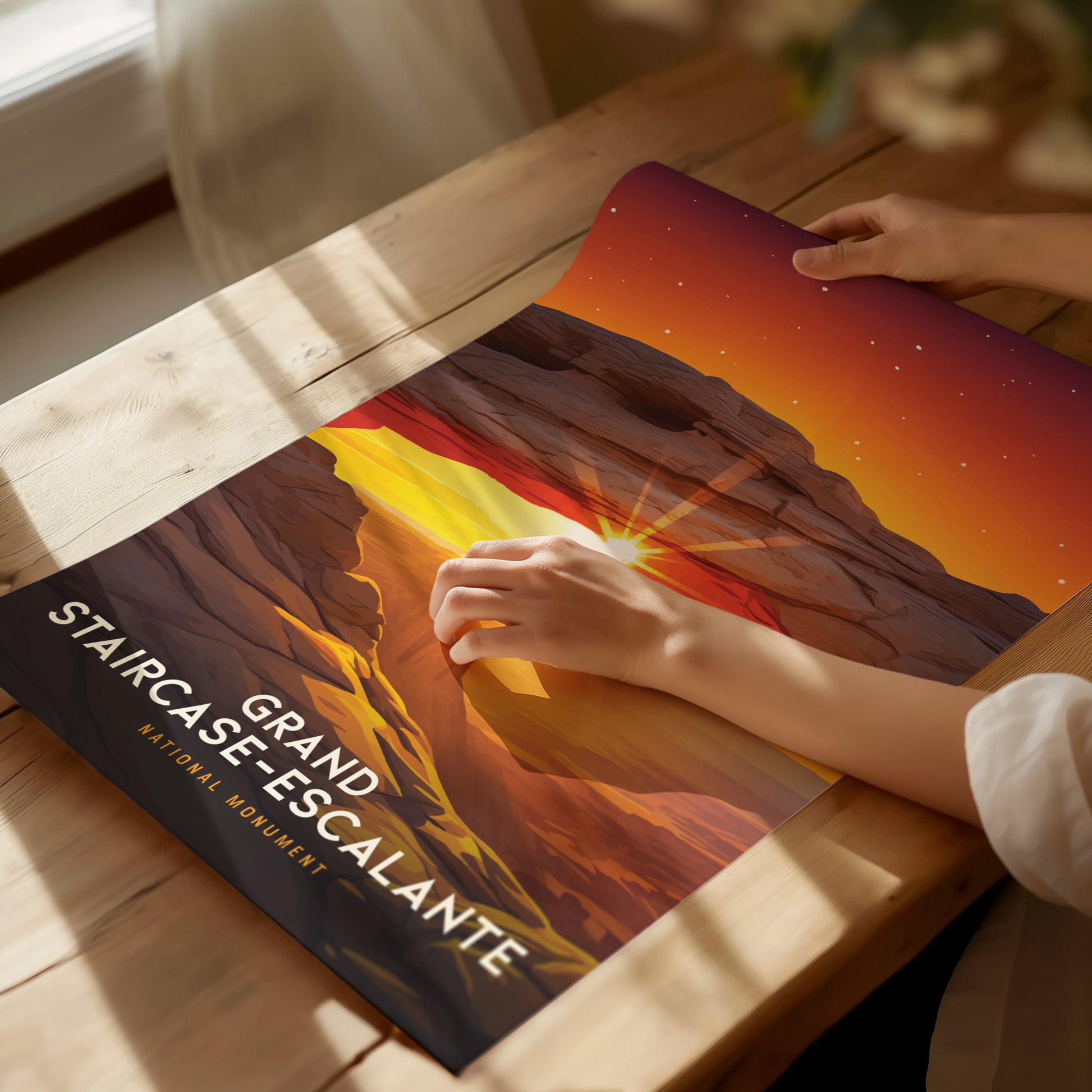 A person unrolls a vibrant Grand Staircase–Escalante poster on a wooden table, showcasing a sunset over rugged rock formations under a starry sky. Sunlight casts soft shadows.