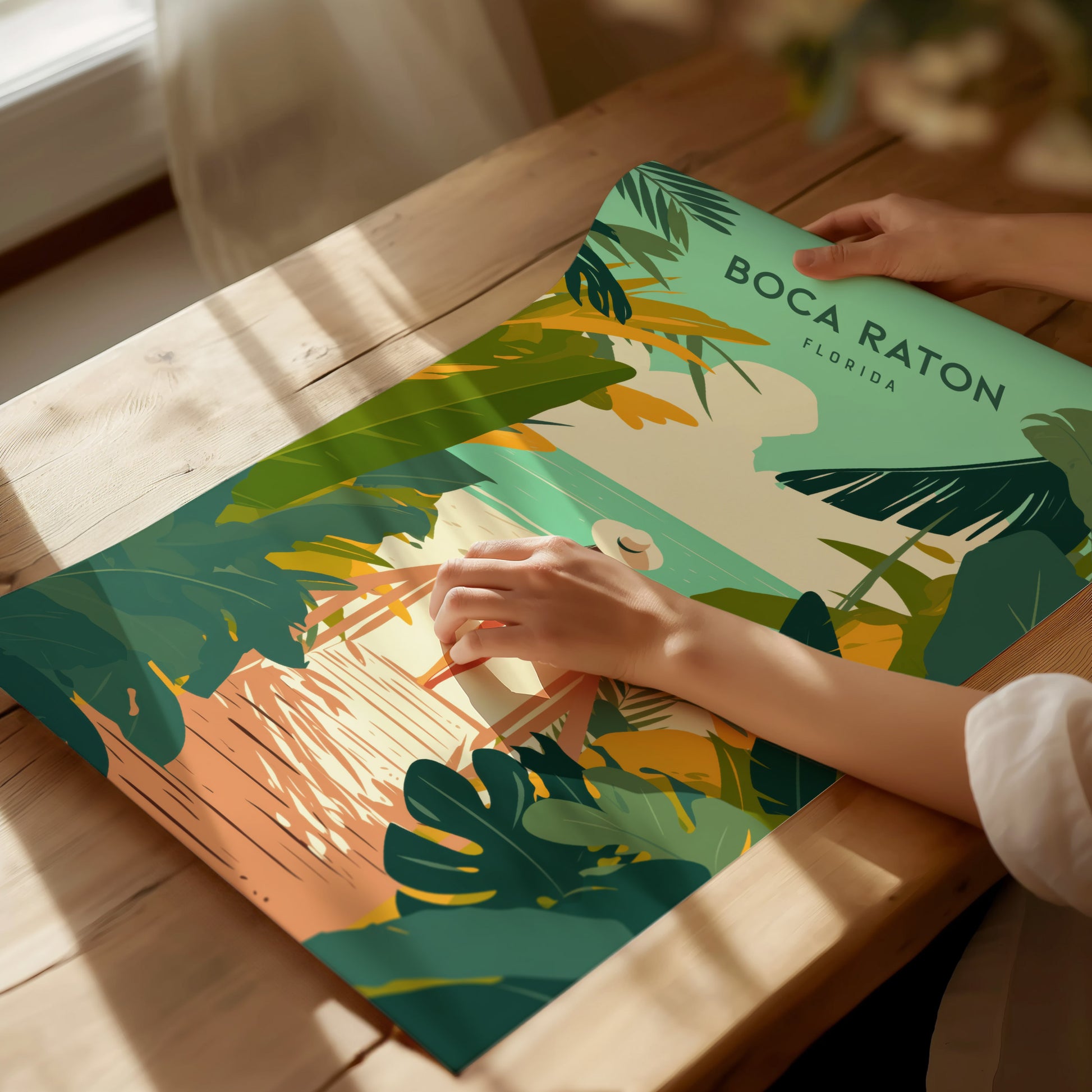 A person arranges a colorful Boca Raton, Florida travel print on a wooden table. The artwork features tropical foliage.