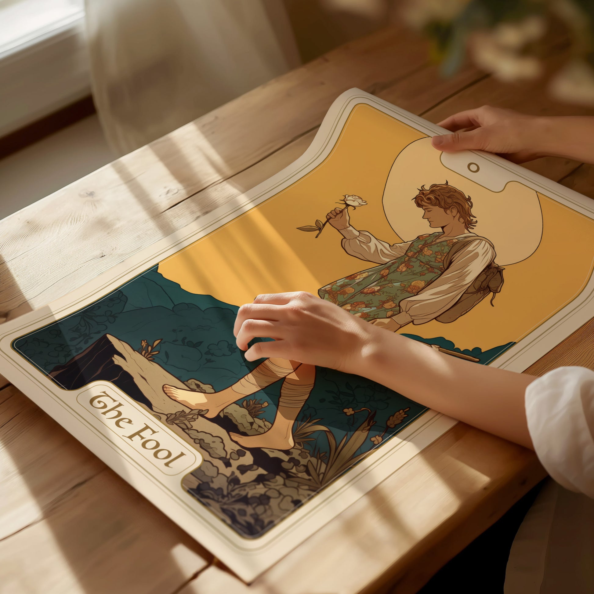 A persons hands hold a tarot poster featuring The Fool, depicting a carefree figure with a white rose, stepping near a cliff, under soft natural lighting.
