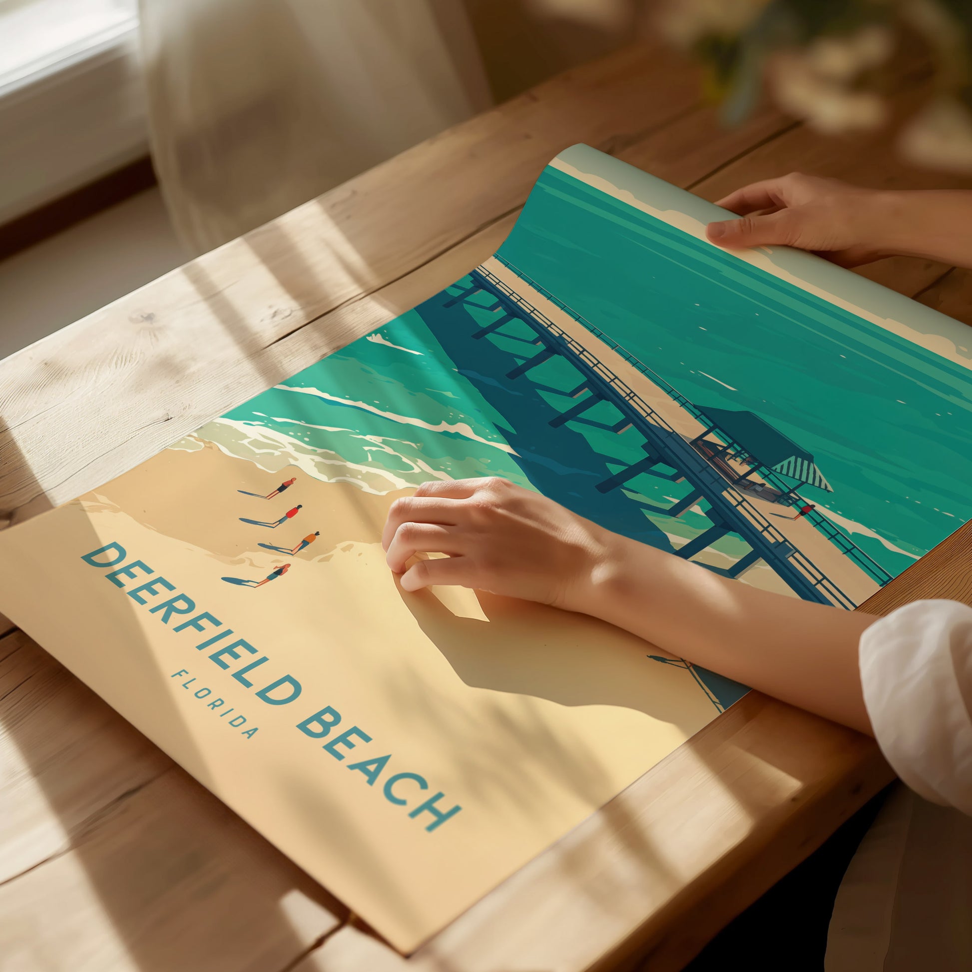 A person unrolls a colorful Deerfield Beach poster on a wooden table, depicting a pier over turquoise waters and sandy beach under a sunny sky.