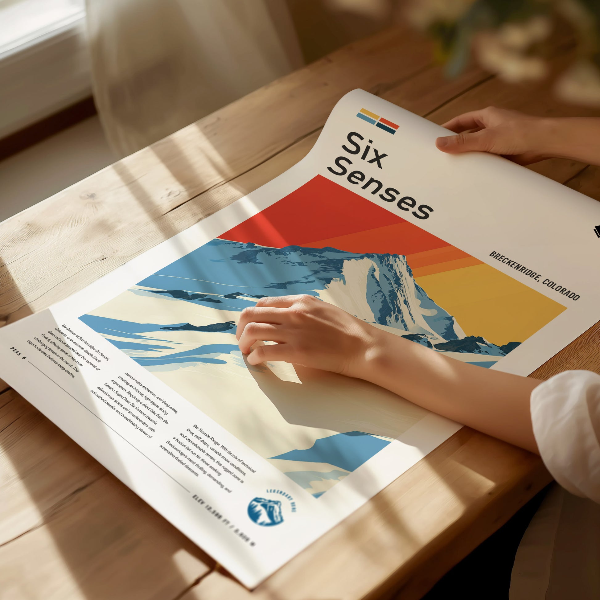 A person examines a colorful poster titled Six Senses depicting a snowy mountain landscape under a vibrant sky on a wooden table. Sunlight casts shadows across the scene.