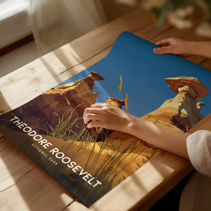 A person examines a vibrant poster of Theodore Roosevelt National Park, featuring hoodoos and grass under a clear blue sky, on a wooden table.