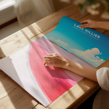 A person unrolls a vibrant Lake Hillier poster on a wooden table, showcasing the pink lake against a blue sky. Sunlight casts soft shadows over the scene.