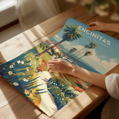 A person unrolls a vibrant Encinitas, San Diego poster featuring palm trees and a coastal landscape on a sunlit wooden table. The artwork exudes a relaxed, beachy vibe.