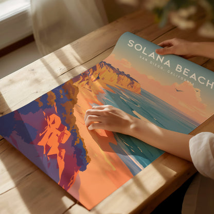 A person is holding a colorful Solana Beach poster on a wooden table, featuring vivid coastal bluffs and ocean waves under a pastel sky.