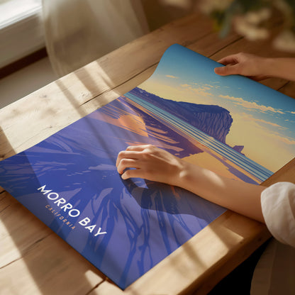 A person is placing a Morro Bay poster on a wooden table. The poster features a stylized depiction of Morro Rock against a colorful sky and ocean backdrop.