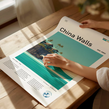 A person adjusts a China Walls poster on a wooden table, depicting turquoise waves and lava rock cliffs with palm trees and coastal homes. Sunlight casts shadows across the scene.