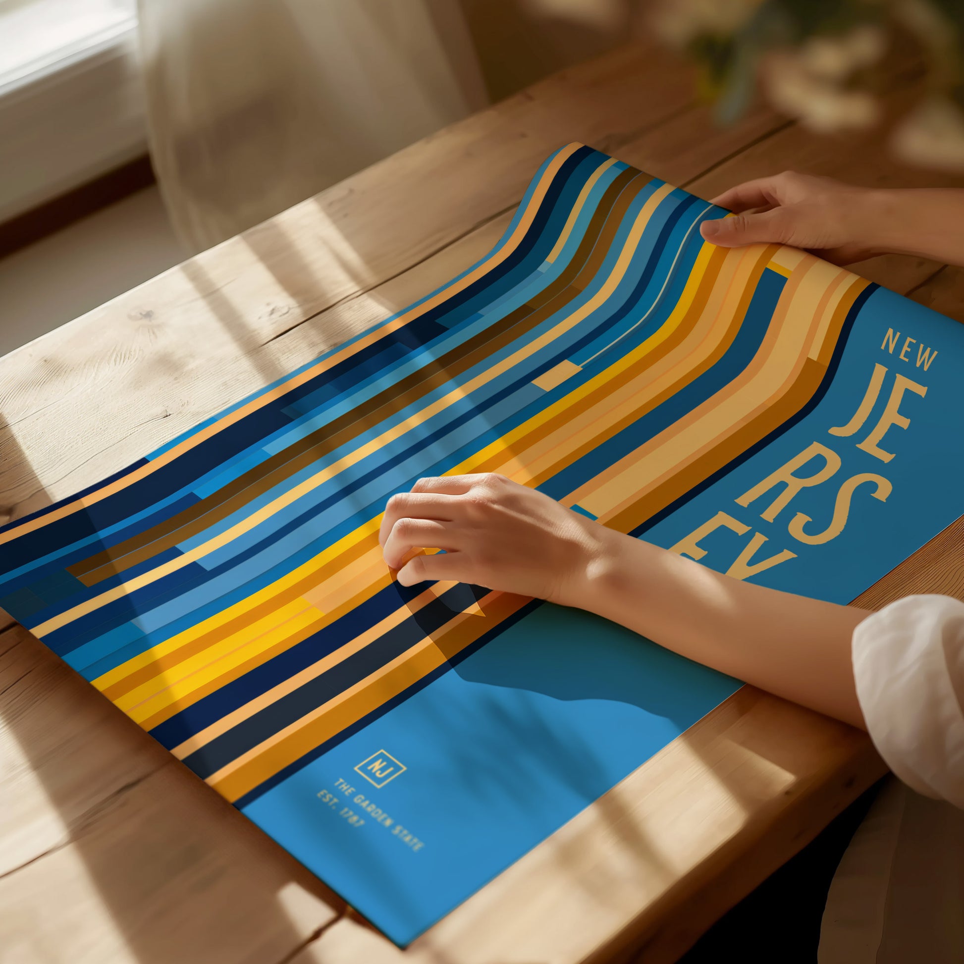 A person unrolls a colorful New Jersey poster with bold stripes and text on a wooden table. Sunlight streams through a nearby window, casting shadows on the surface.