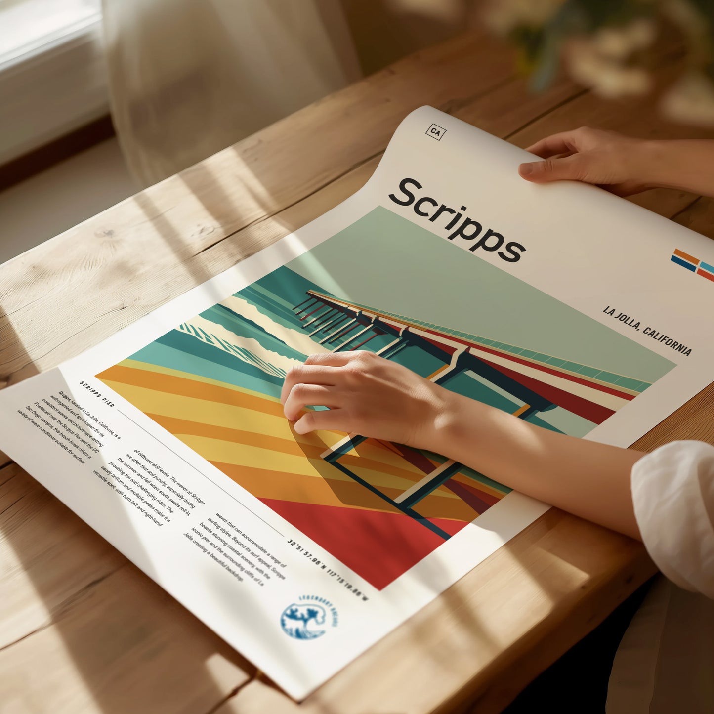 A person holds a colorful poster depicting Scripps Pier in La Jolla, California, on a wooden table. The artwork features vibrant coastal hues and bold architectural lines.