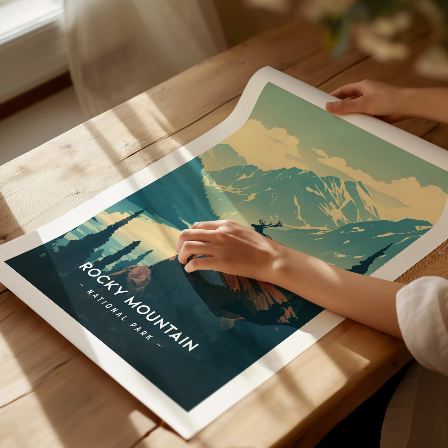 A person is gently unrolling a Rocky Mountain National Park poster on a wooden table, showcasing a minimalist landscape with mountains and trees. Sunlight casts soft shadows across the scene.