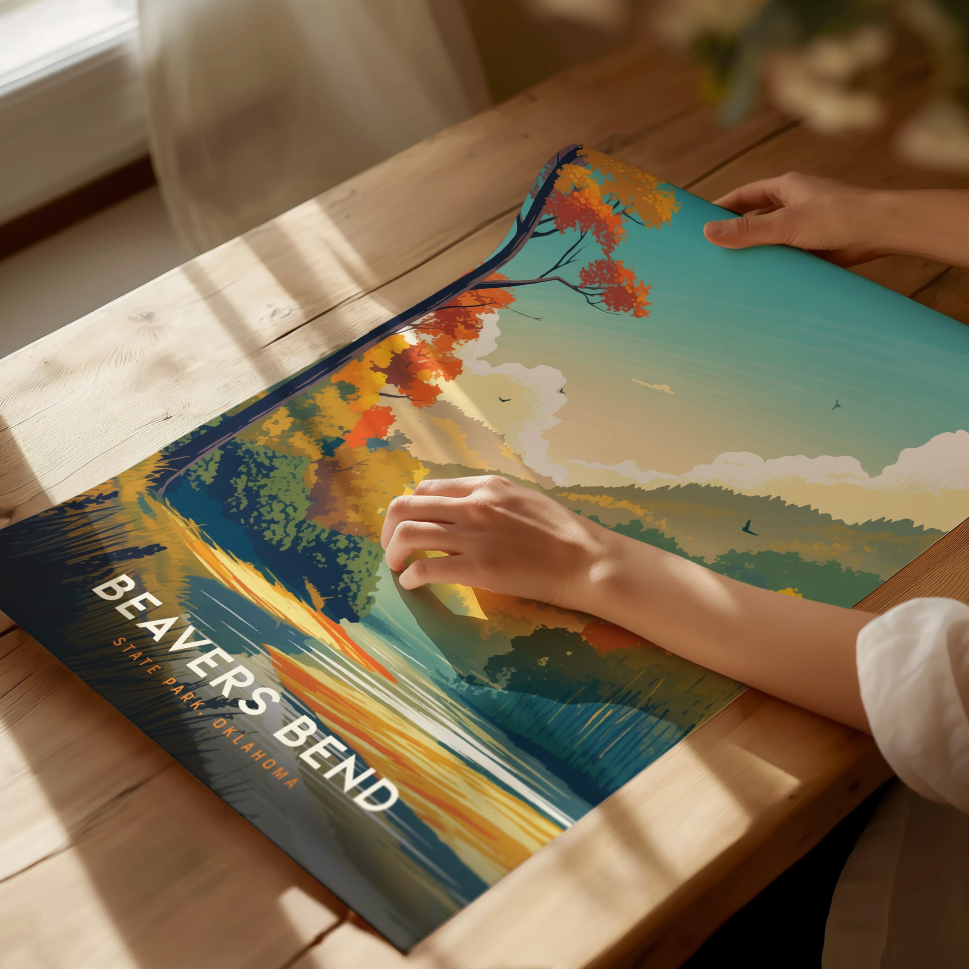 A person adjusts a colorful Beavers Bend State Park poster on a wooden table, depicting vibrant autumn trees and a serene lake. Sunlight softly illuminates the scene.
