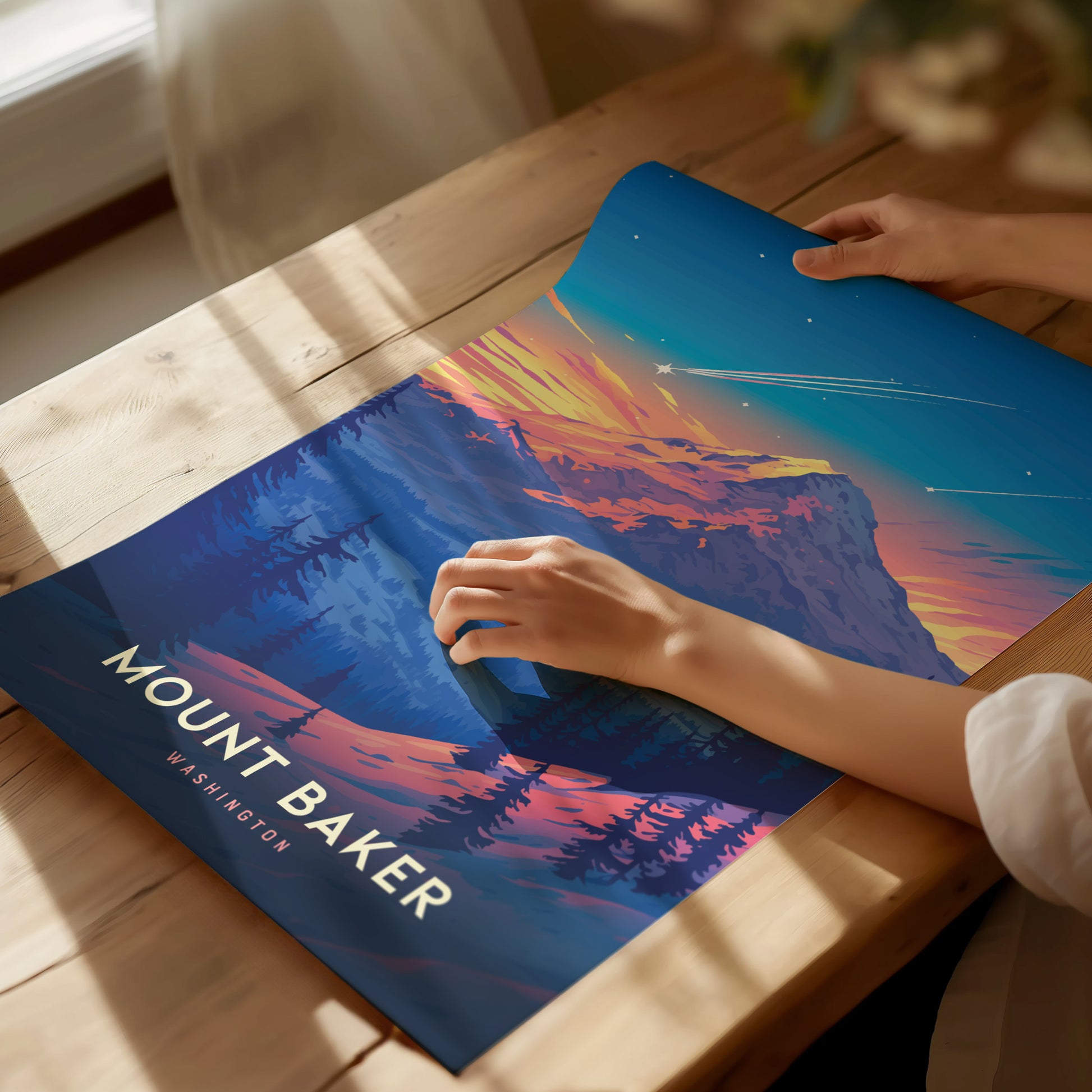 A person is holding a vibrant Mount Baker poster featuring a colorful sunset and forested landscape. The artwork lies on a wooden table illuminated by soft natural light.