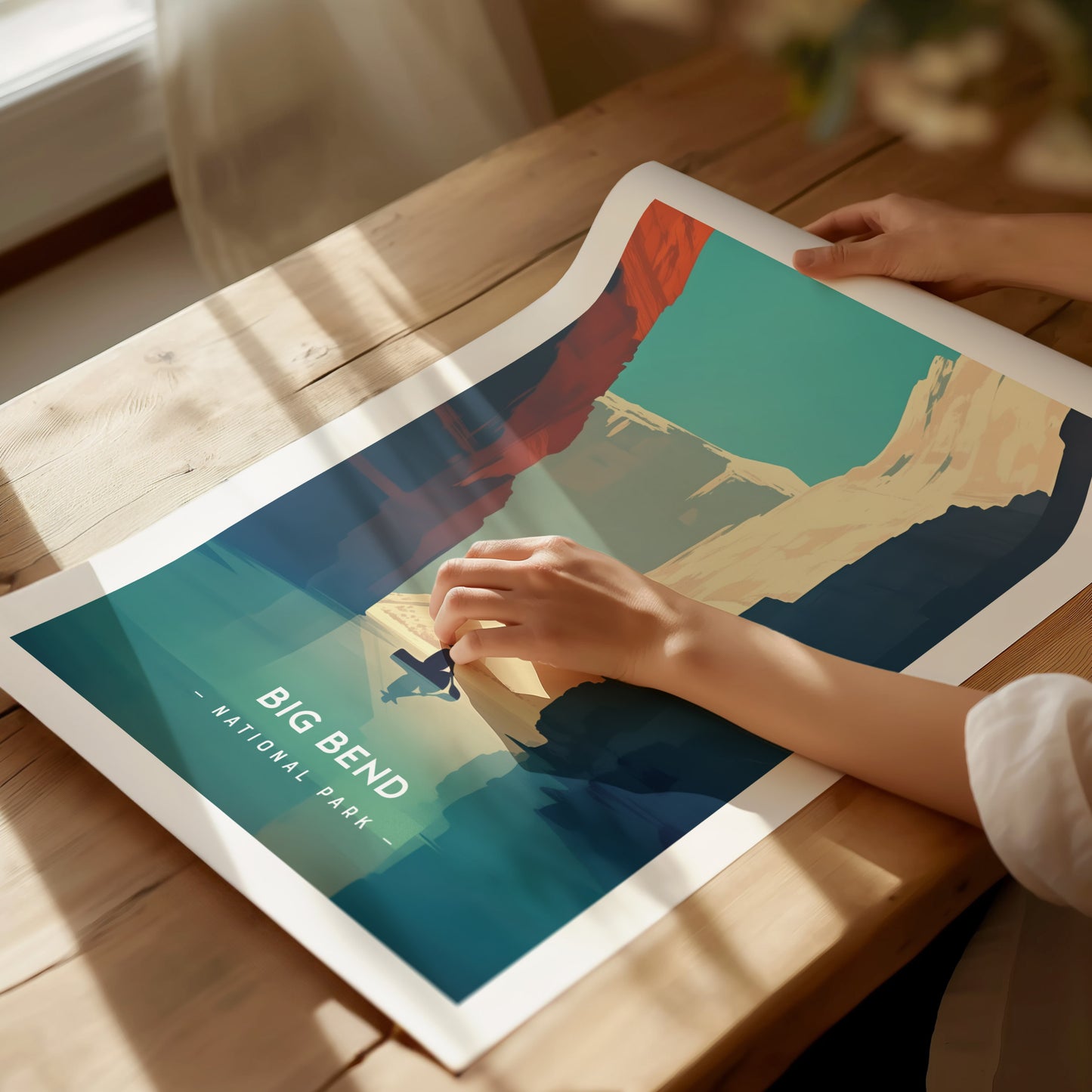 A person gently holds a vibrant, minimalist Big Bend National Park poster on a wooden table. The artwork features colorful desert landscapes under a clear blue sky.