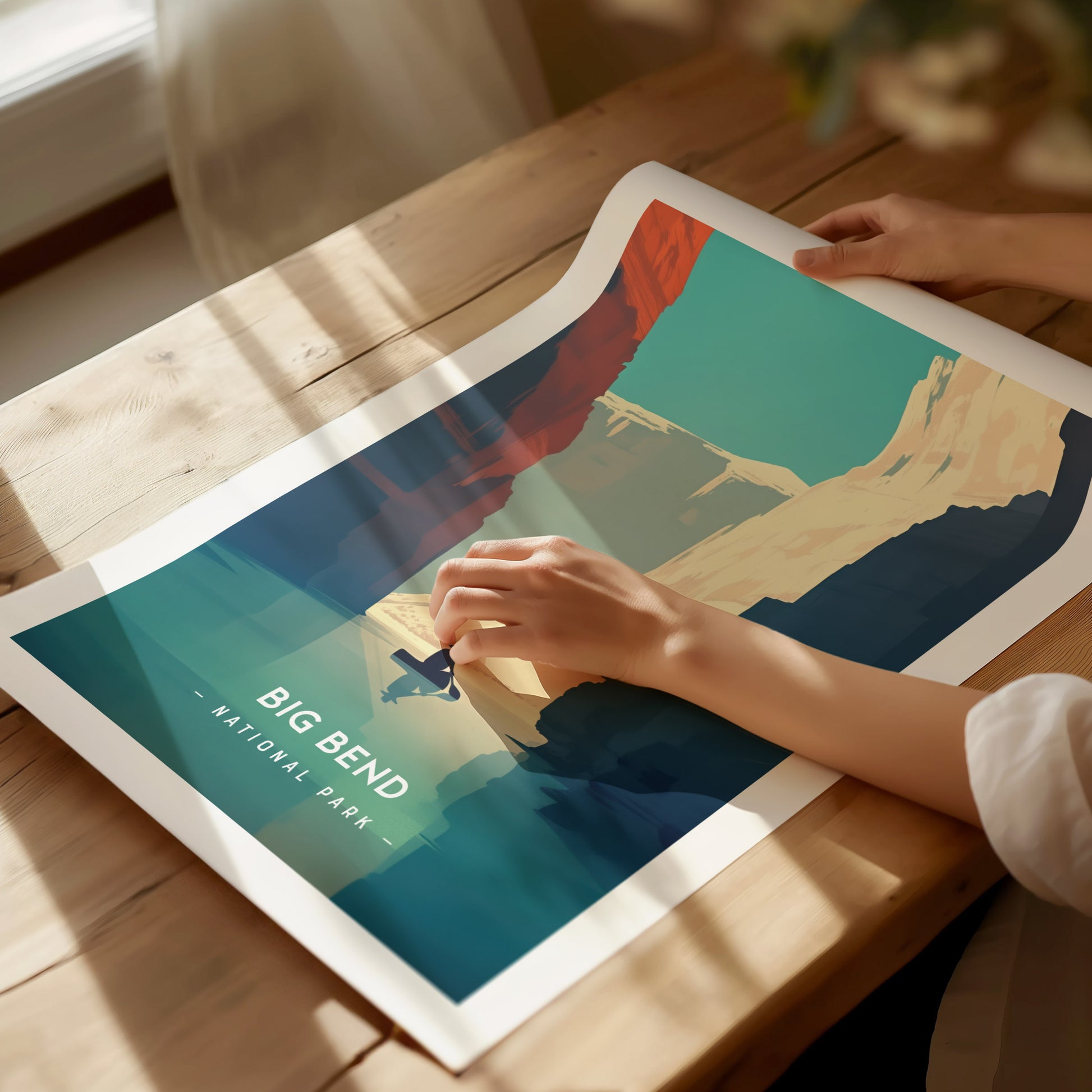 A person gently holds a vibrant, minimalist Big Bend National Park poster on a wooden table. The artwork features colorful desert landscapes under a clear blue sky.