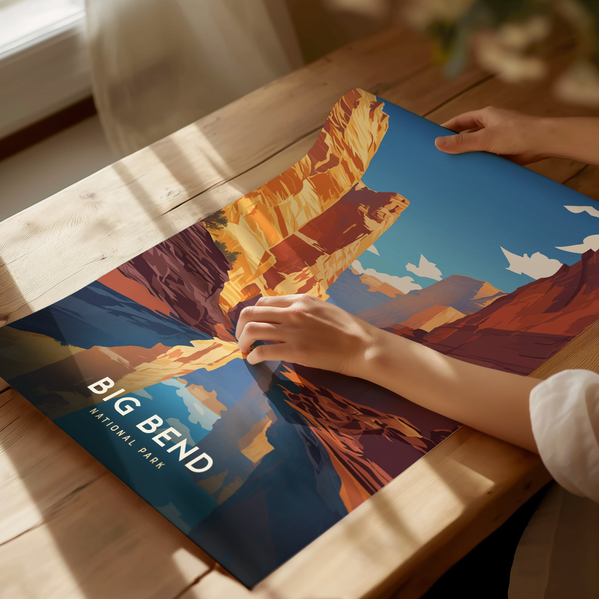 A person holds a colorful poster of Big Bend National Park on a wooden table, showcasing vivid desert landscapes under a bright blue sky.